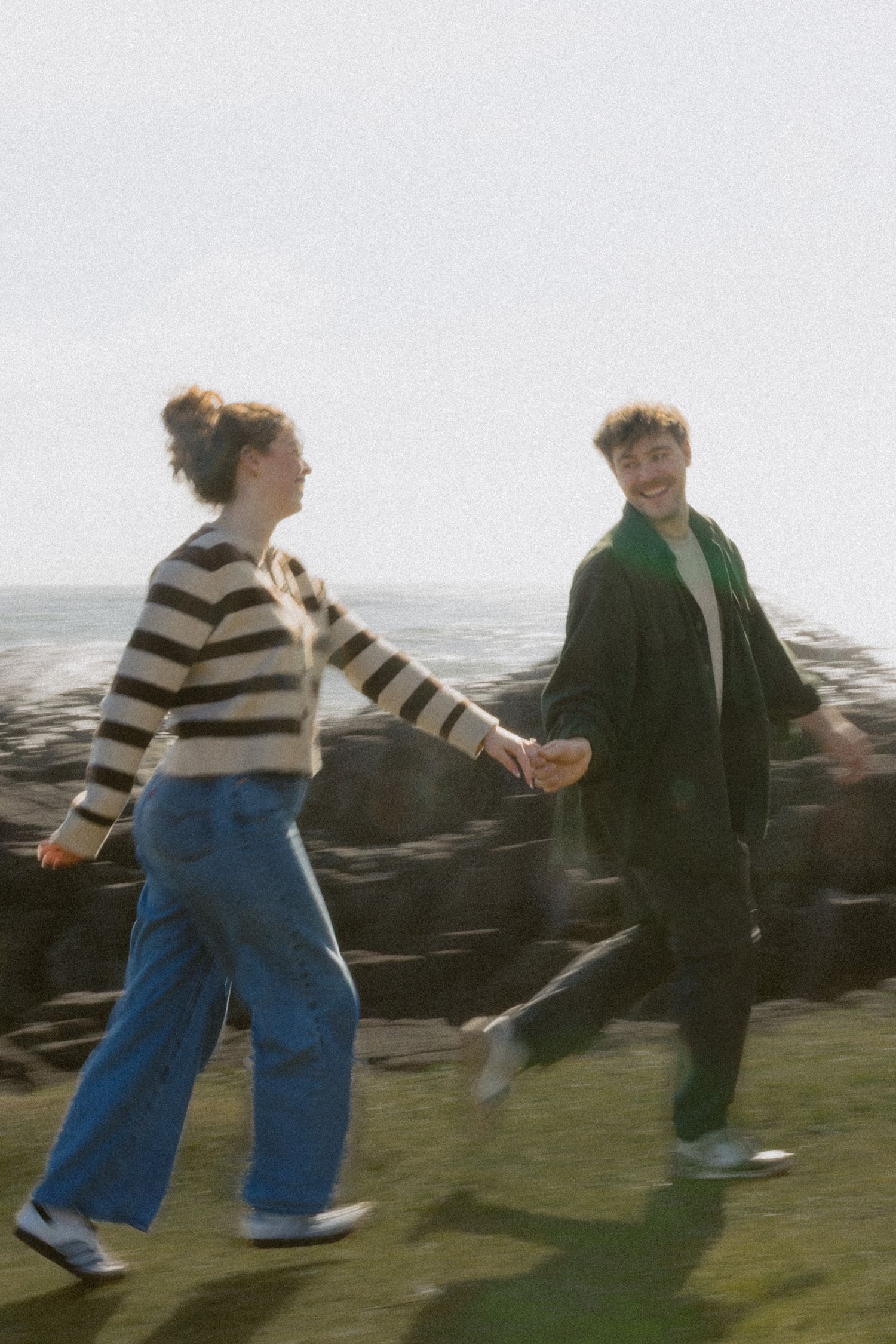 Happy engaged couple holding hands and walking on grassy coastal bluff in warm sunlight.
