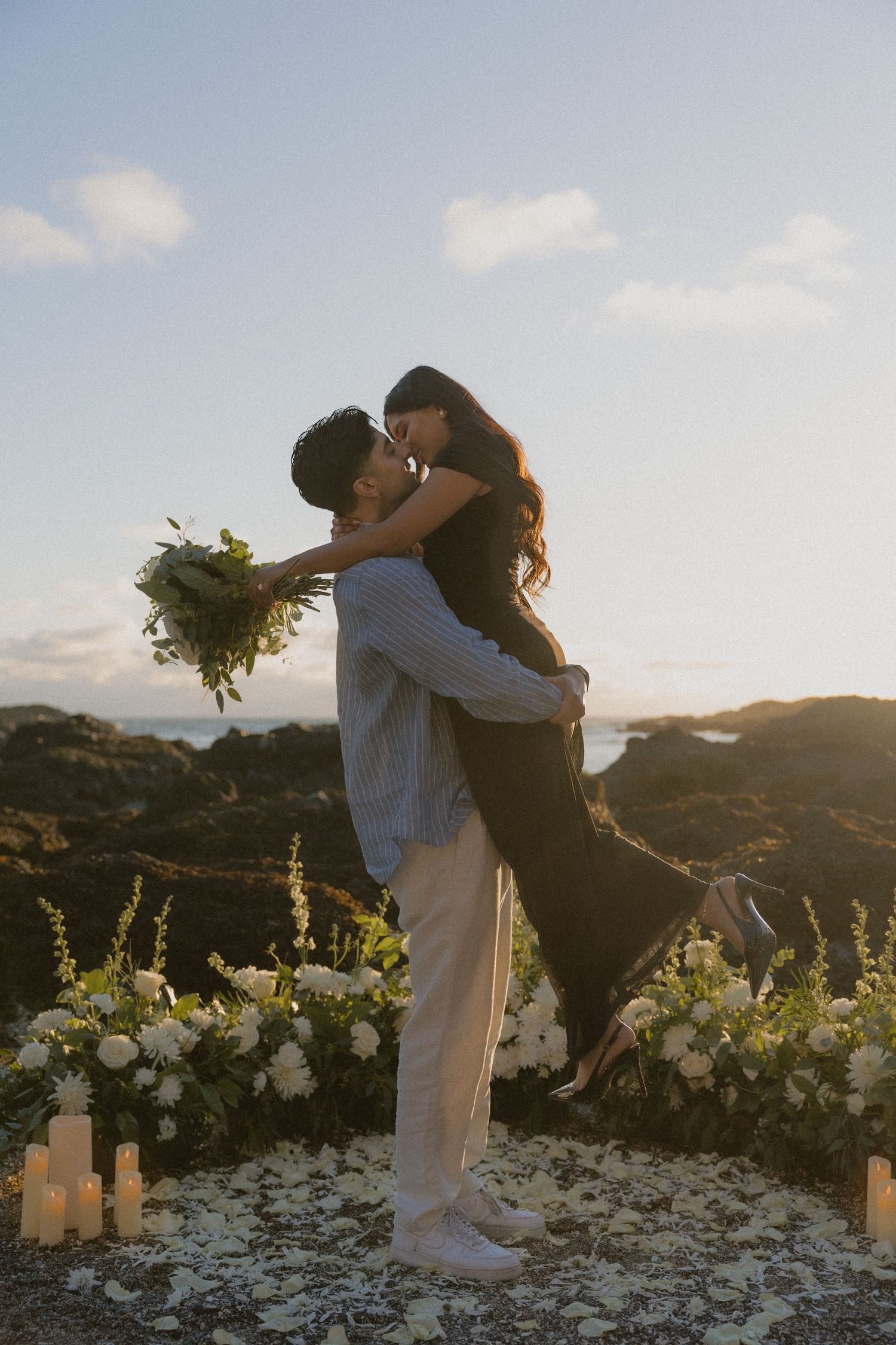 Couple sharing a romantic kiss at sunset on rocky coast, surrounded by white flowers and candles.