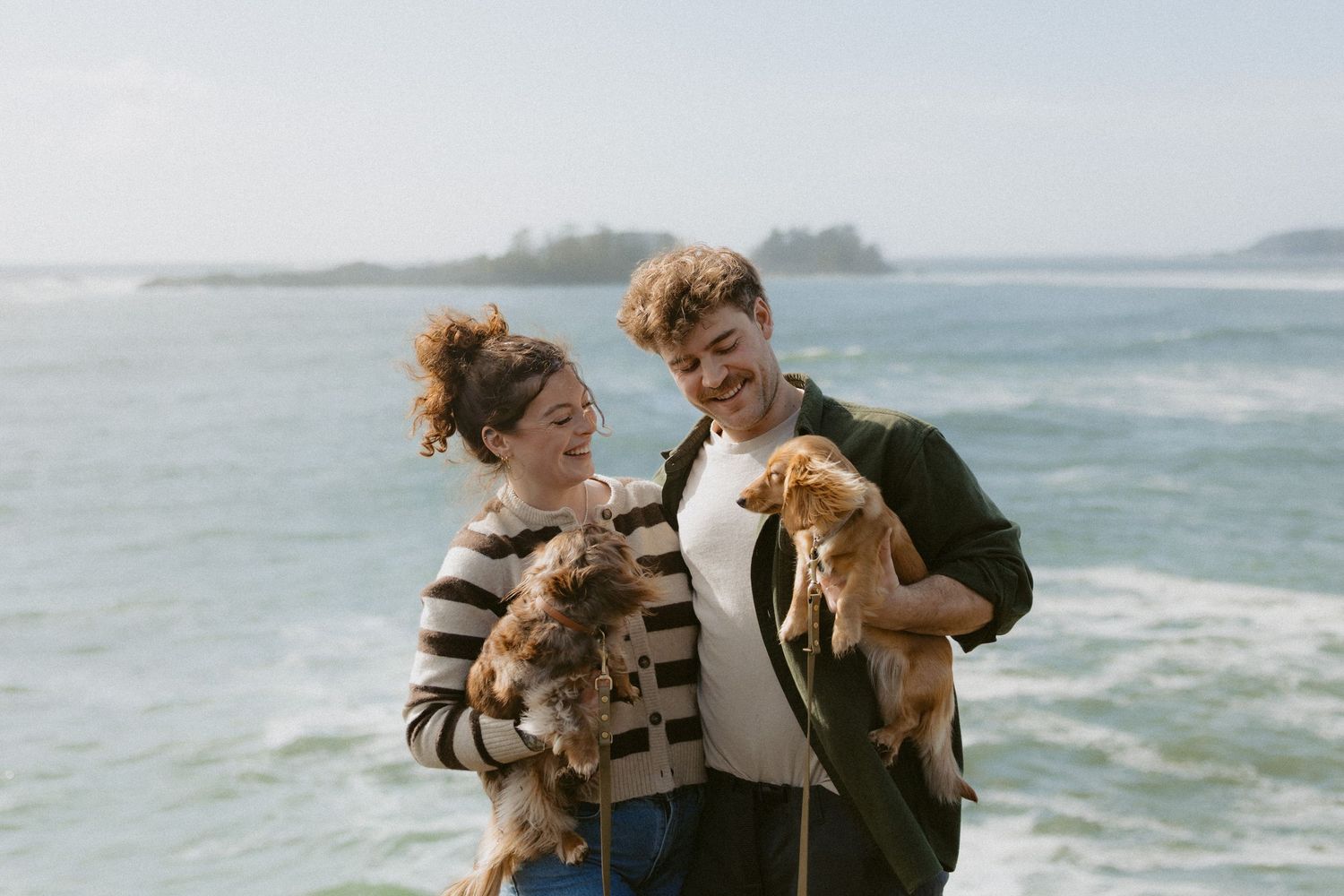 Happy couple holding small dogs while smiling at each other on a scenic ocean beach.