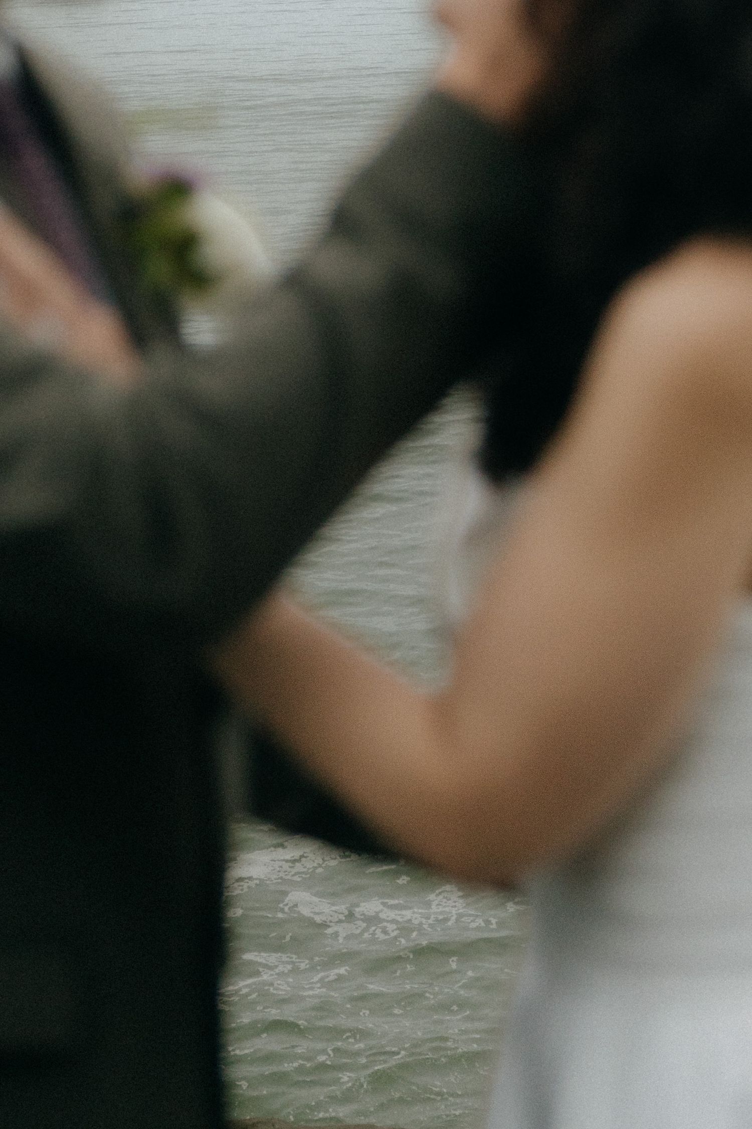 Blurred romantic photo of a couple in wedding attire embracing near water, groom in suit with boutonniere.