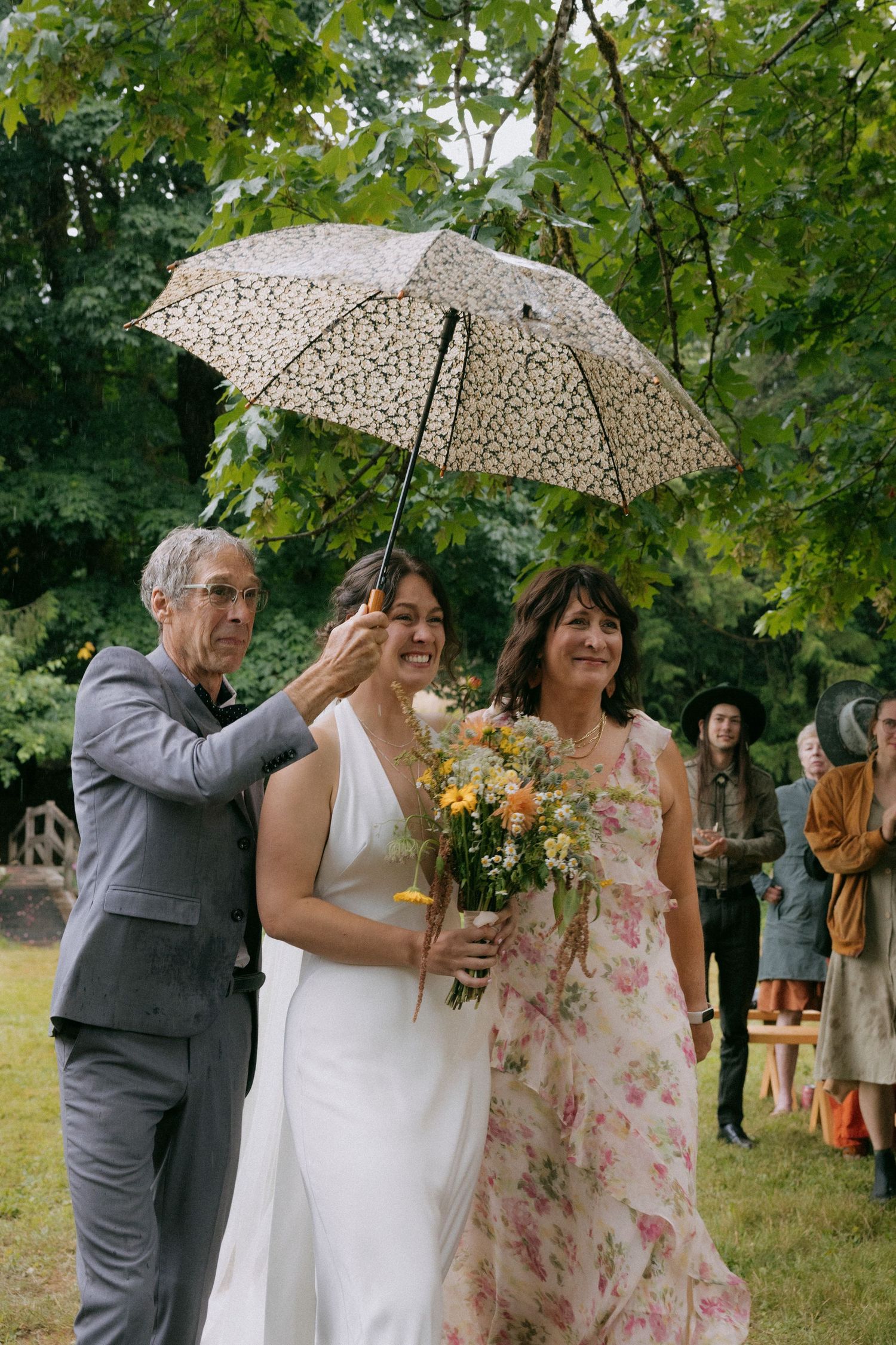 Bride walks down outdoor aisle with parents holding umbrella, carrying wildflower bouquet in white gown.
