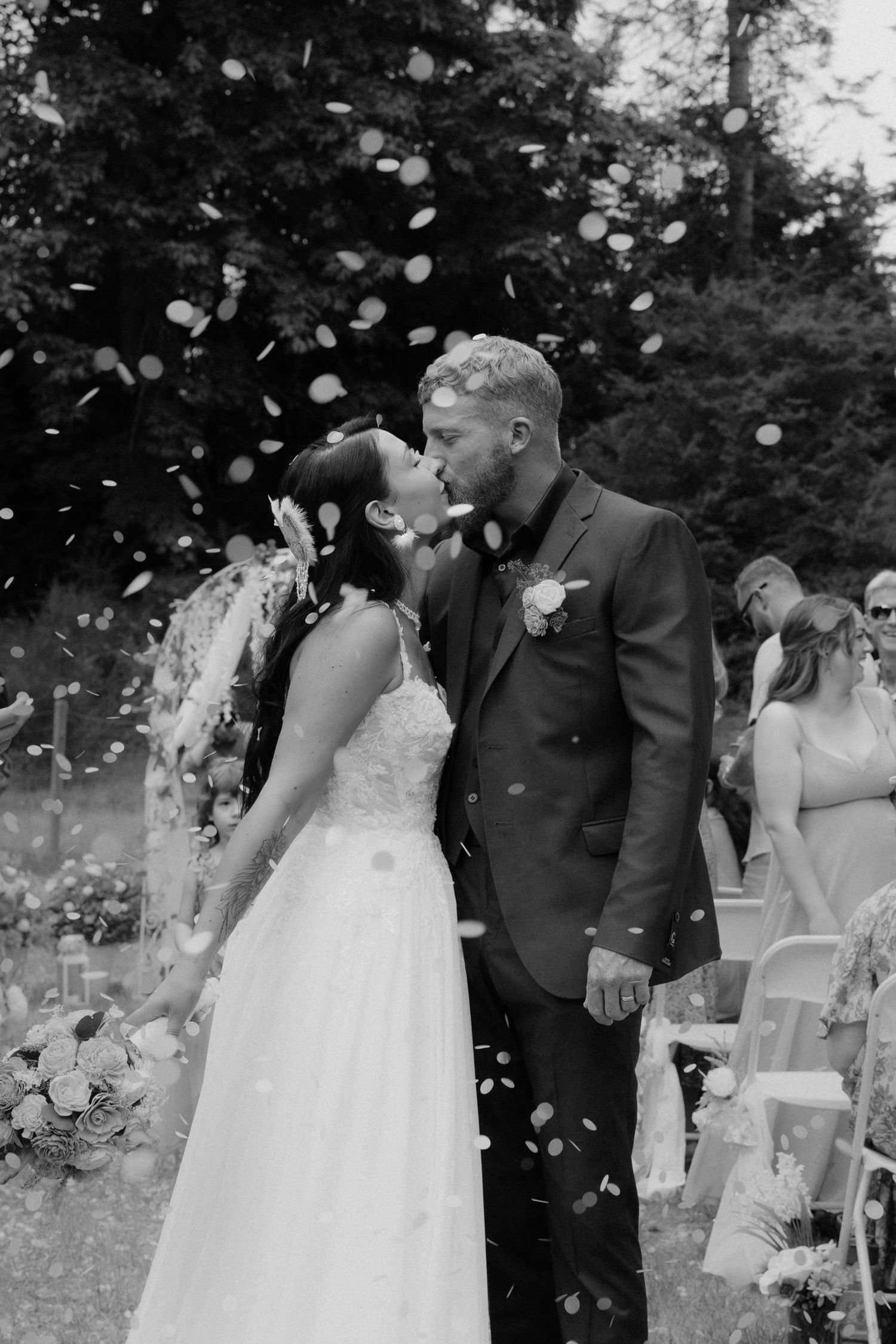 Black and white photo of a bride and groom sharing a kiss as confetti falls around them at an outdoor wedding ceremony.