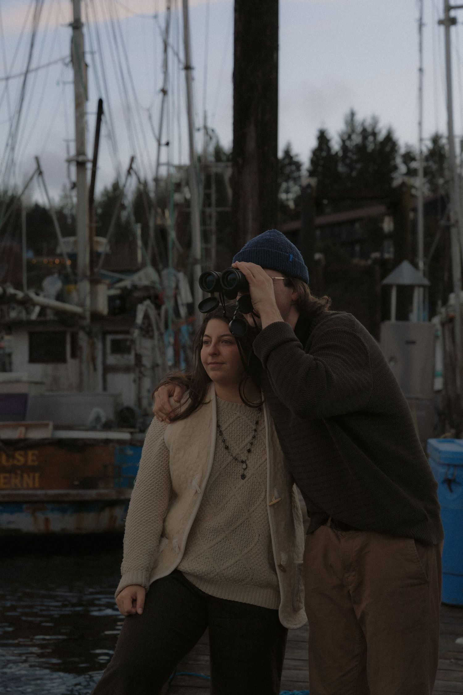 Tofino lifestyle couple looking through binoculars together at a Tofino marina during sunset surrounded by boats