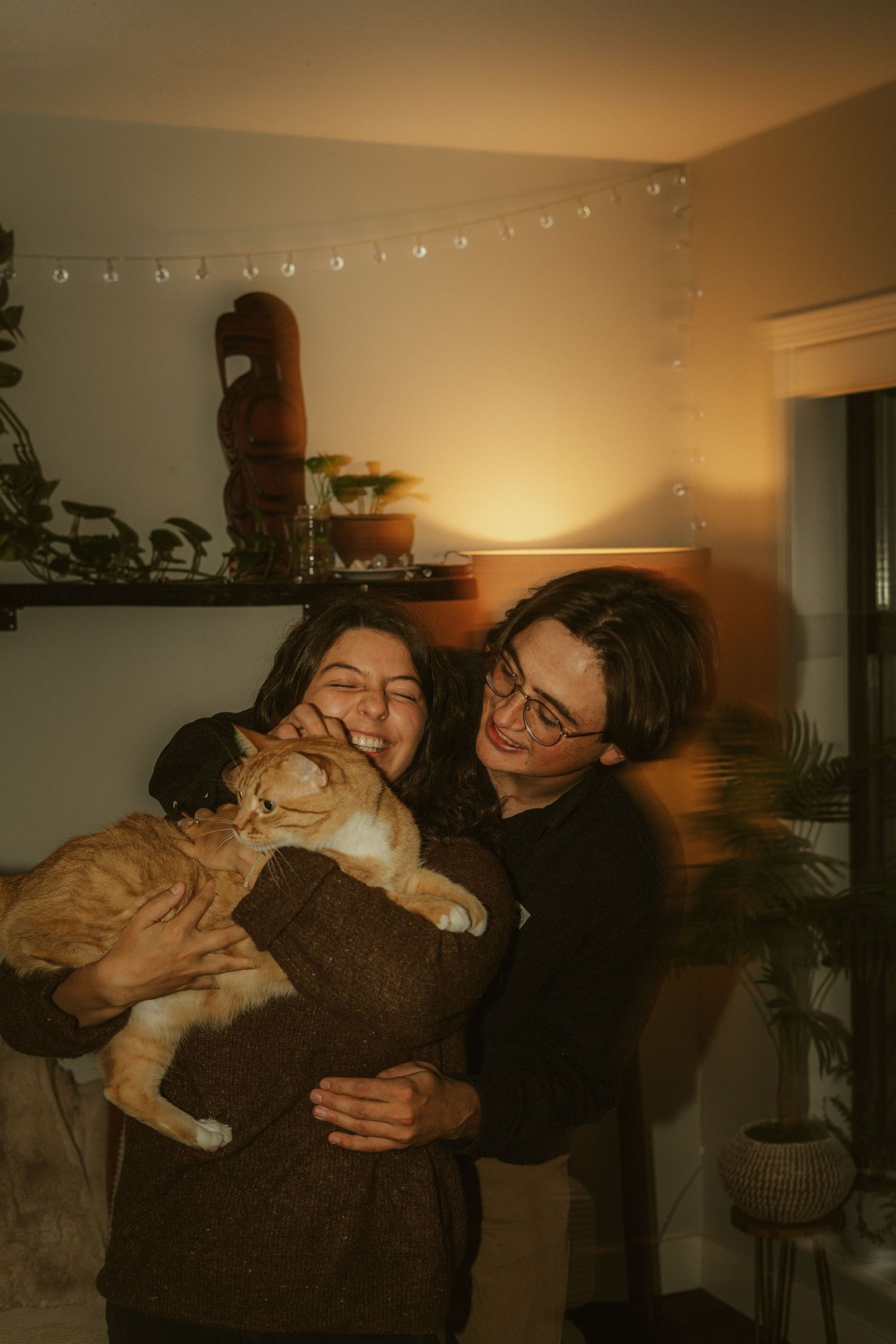 Tofino couple cuddling with orange cat under warm string lights, Mariana and partner