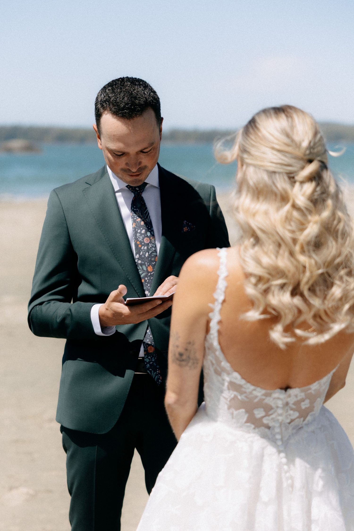 Groom in green suit reads vows from card to blonde bride in lace wedding dress on sunny beach.