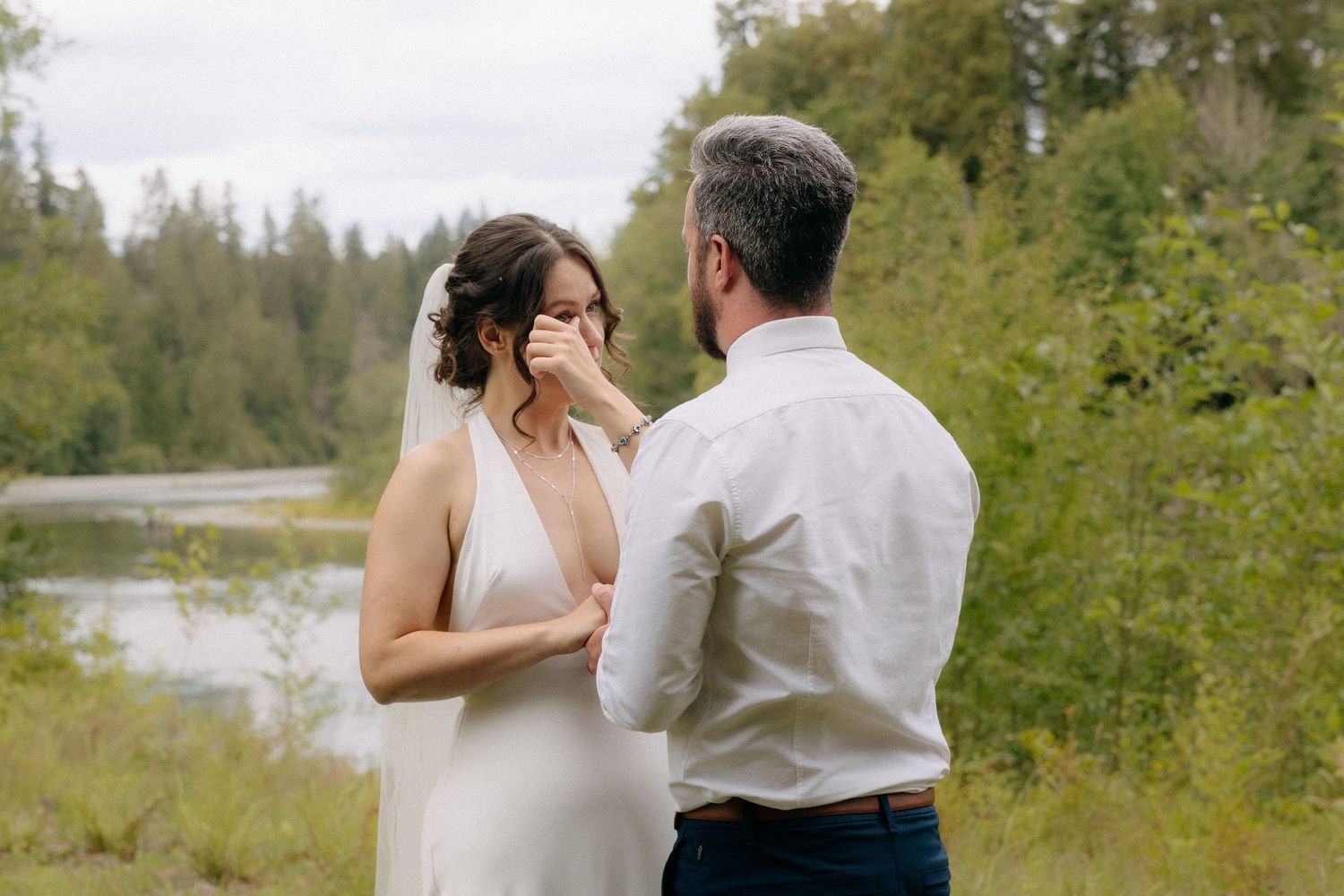 Emotional bride wiping tears during first look with groom outdoors near river surrounded by lush green trees.