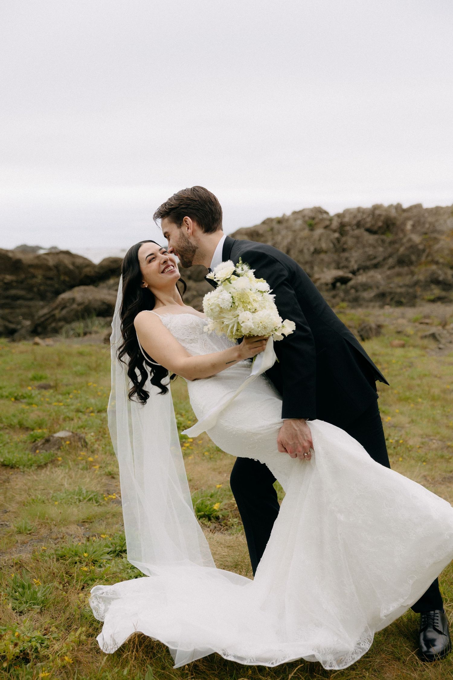 Groom in black suit kisses bride in white gown holding white bouquet outdoors near rocky coastal landscape.