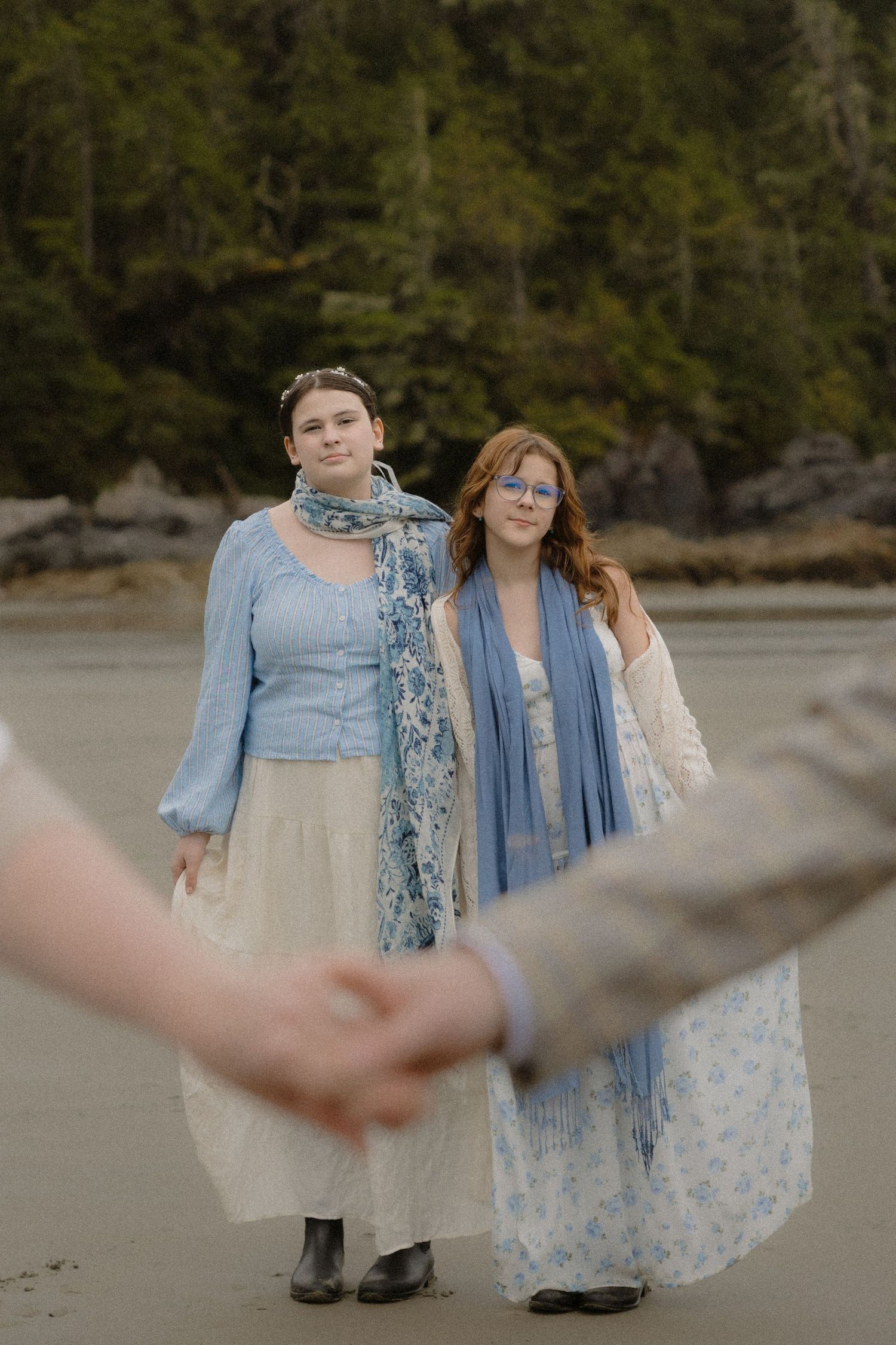 Two people in flowing blue dresses stand together on a misty beach with rocky cliffs.