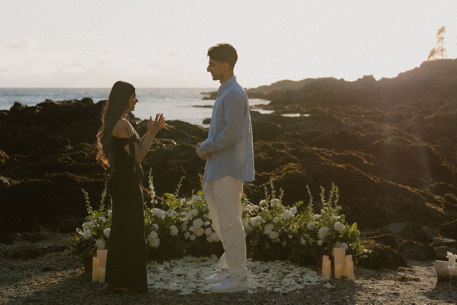 Couple exchanging vows at a romantic beach proposal with white floral arrangements and candles at golden hour.