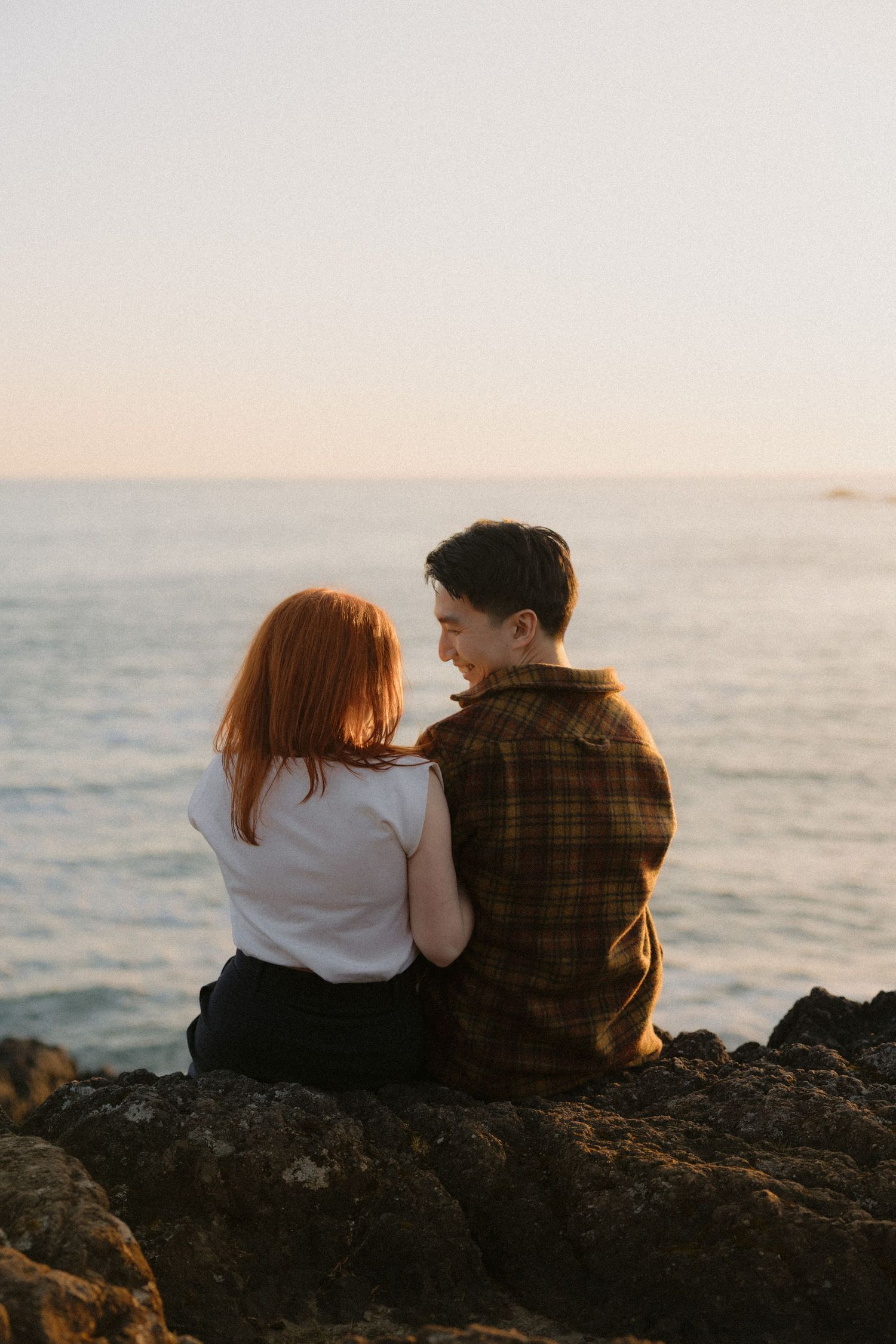 Couple sitting together on rocks overlooking calm ocean at golden hour sunset.