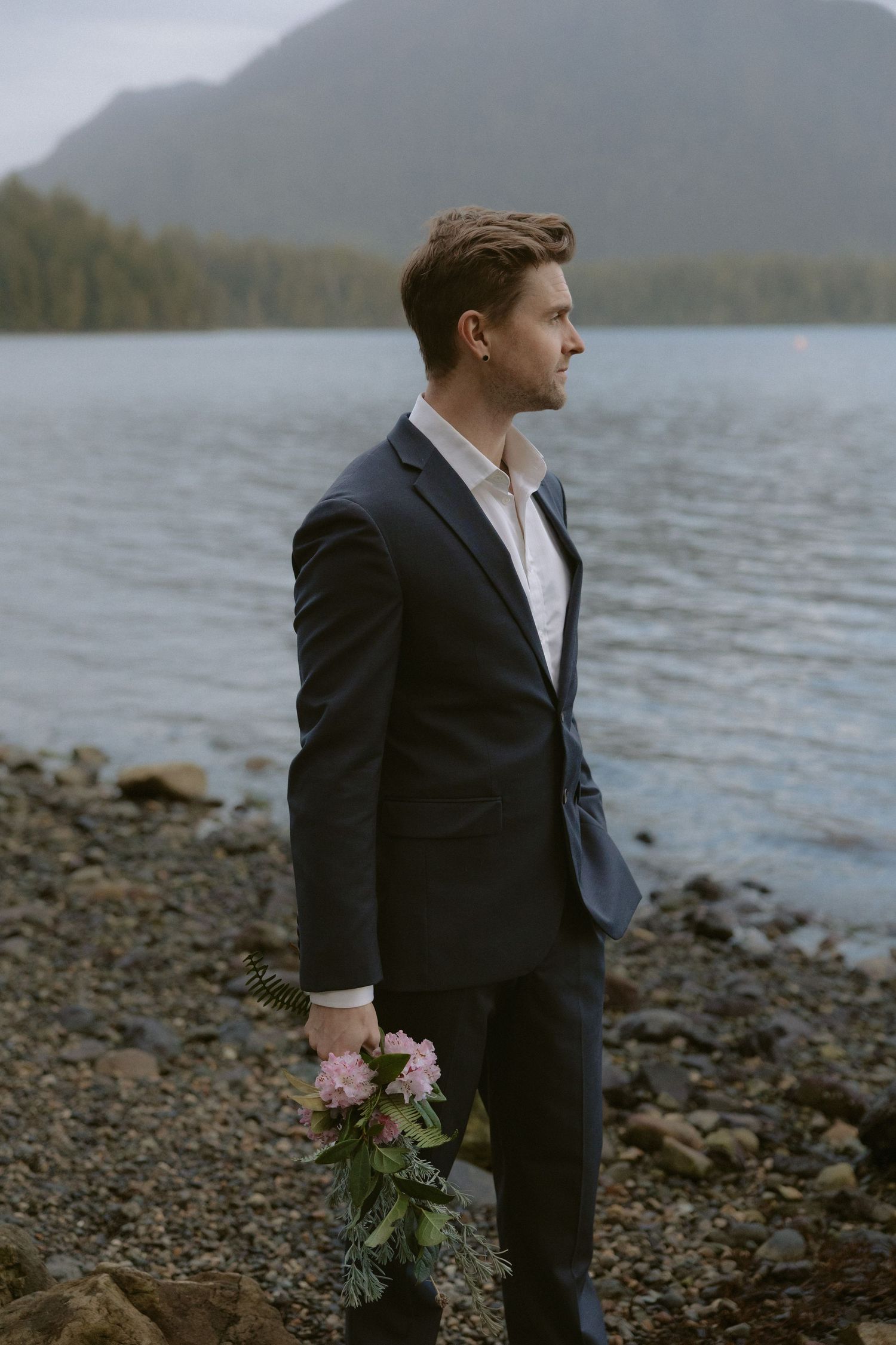 Ucluelet groom in dark suit holding flowers stands oceanside in the Pacific Rim with mountains in background