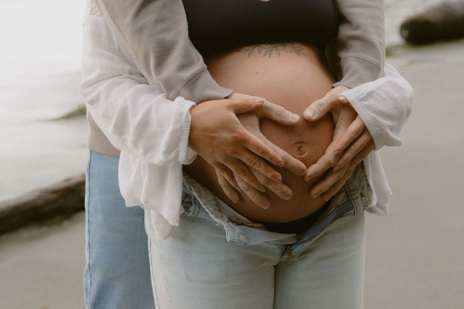 Hands forming heart shape over pregnant belly during beach photoshoot.