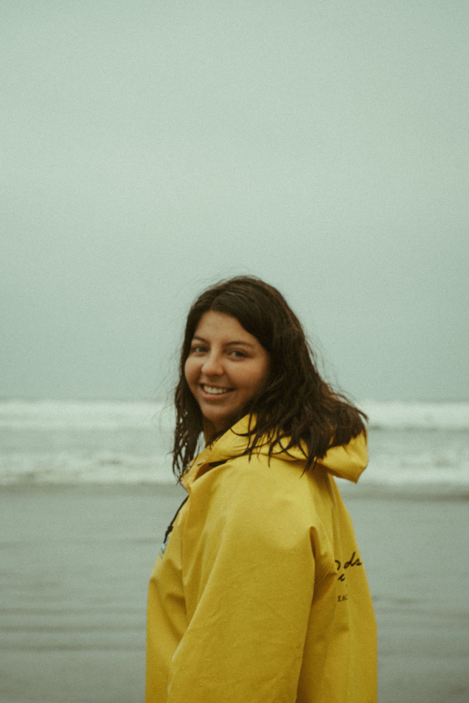 Tofino wedding photographer Mariana in a bright yellow raincoat stands against a misty coastal backdrop on Cox Bay Tofino
