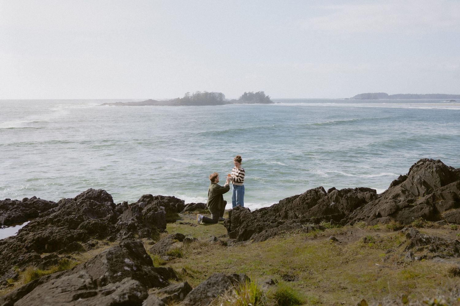 Couple holding hands on rocky coastal shoreline with ocean waves and small island in background.
