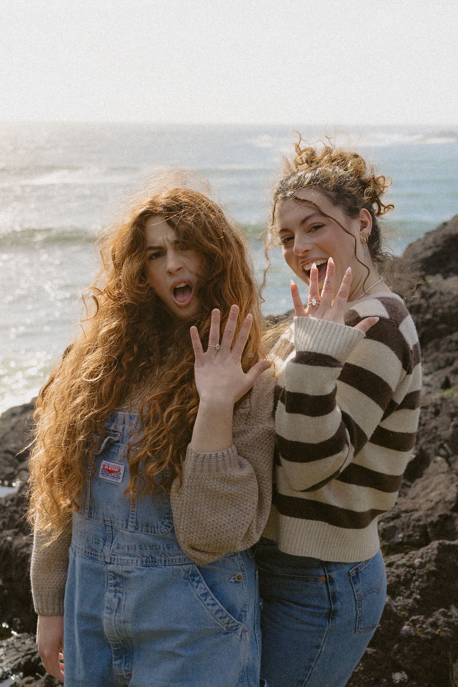 Two excited women showing off engagement ring on windy rocky beach with ocean backdrop.