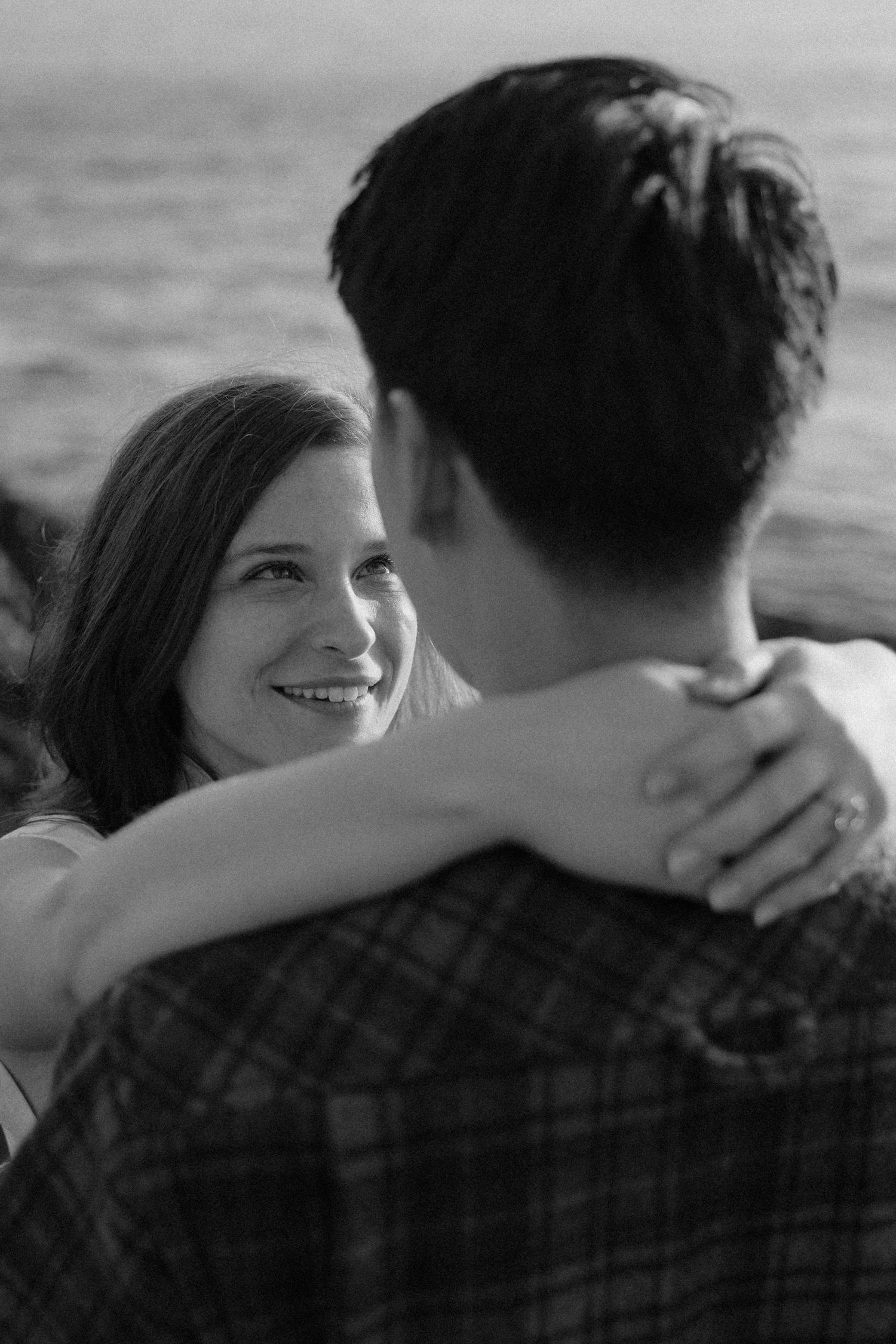 Smiling woman embracing man in plaid shirt during romantic black and white couples photography session.