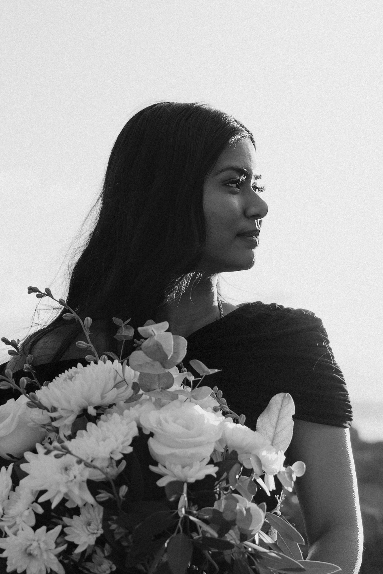 Black and white portrait of a young woman holding a bouquet of roses and daisies, gazing thoughtfully to the side.