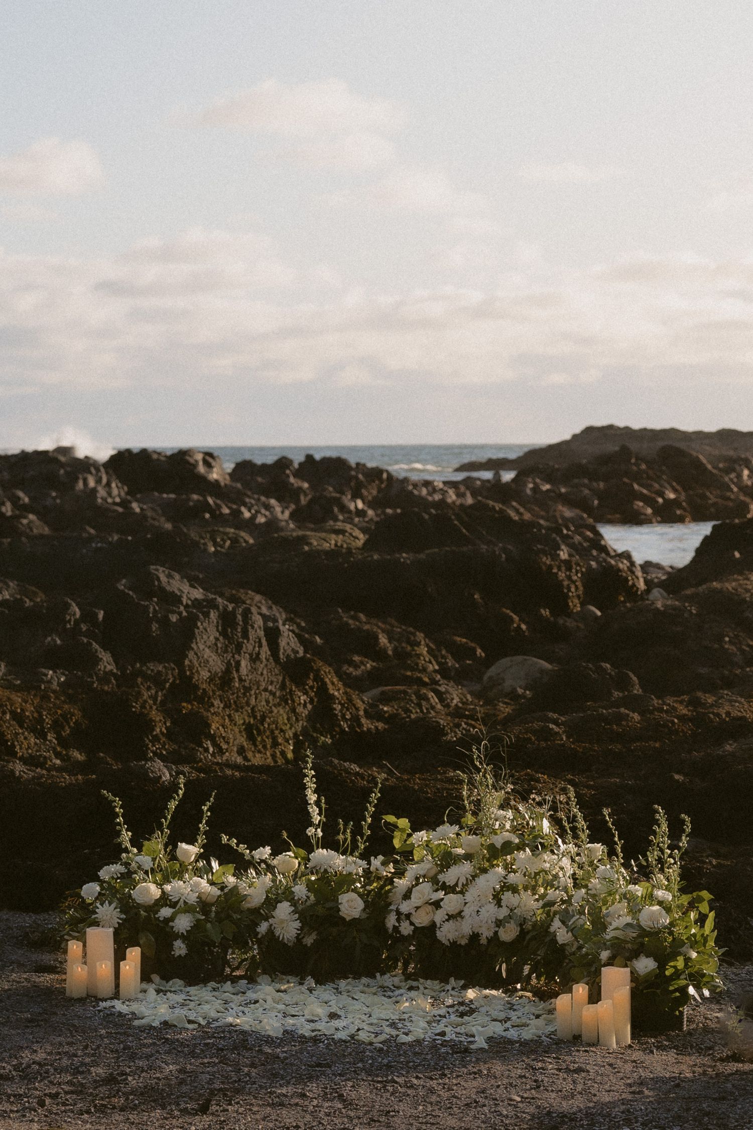 Romantic coastal wedding altar with white flowers and candles on volcanic rocks by the ocean.