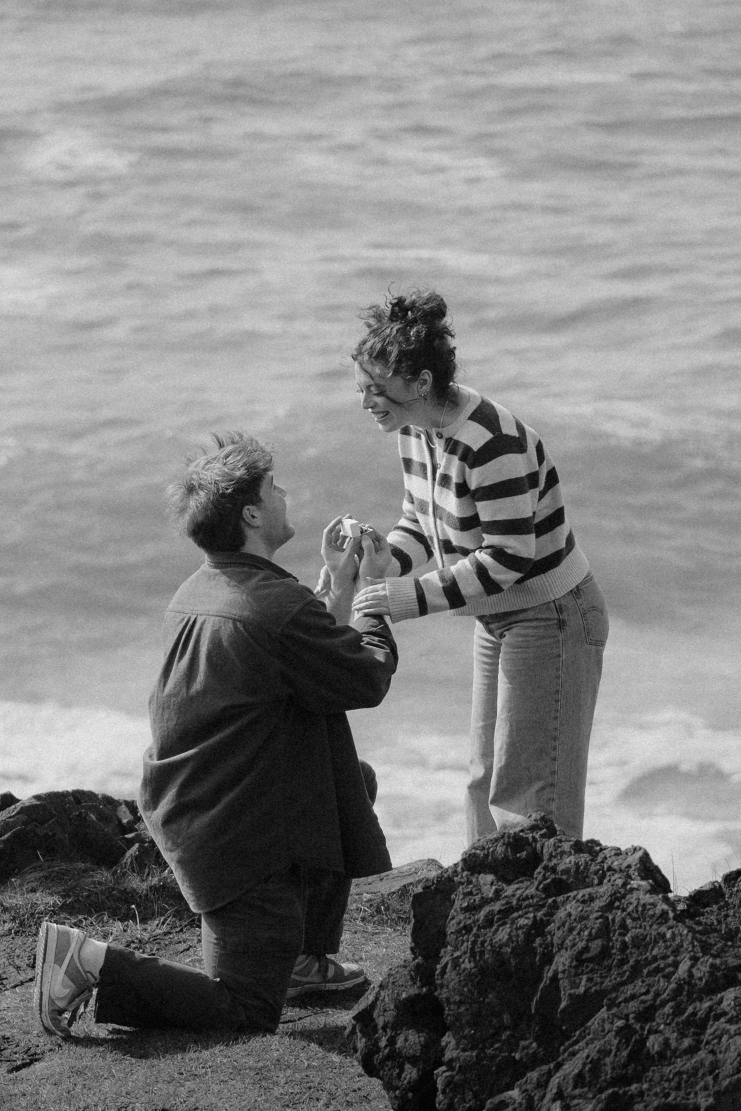 Man proposing on one knee to woman on rocky oceanside cliffs in black and white photo.
