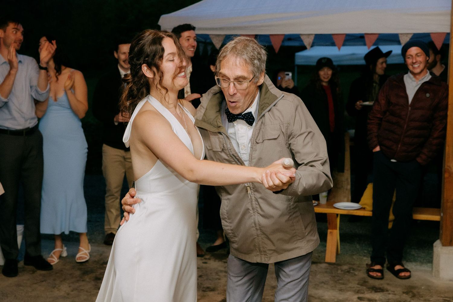 Bride in white gown dances with elderly man in bow tie at outdoor wedding reception with guests applauding.