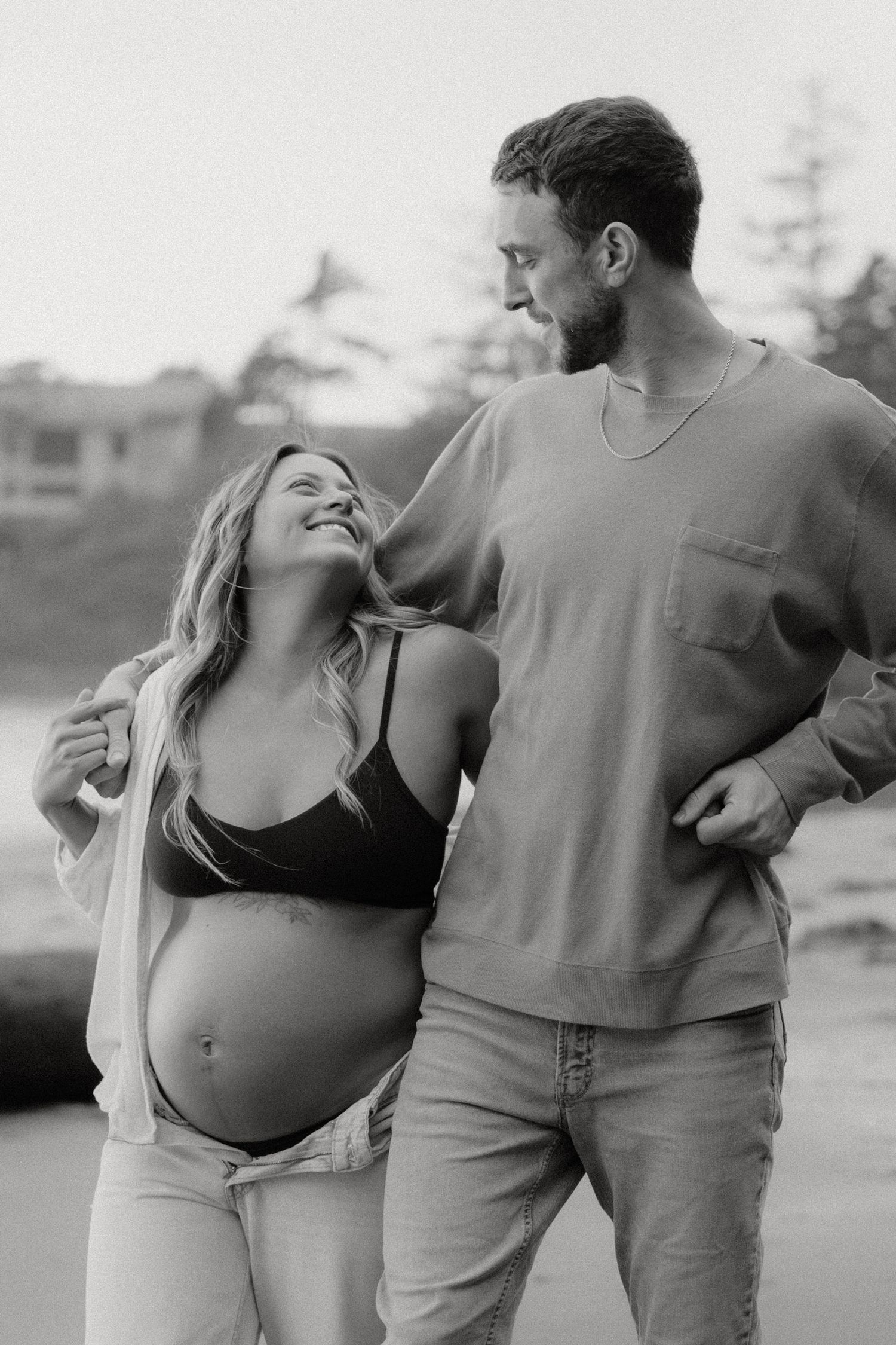 Black and white maternity photos capture intimate moments between a couple on the beach.