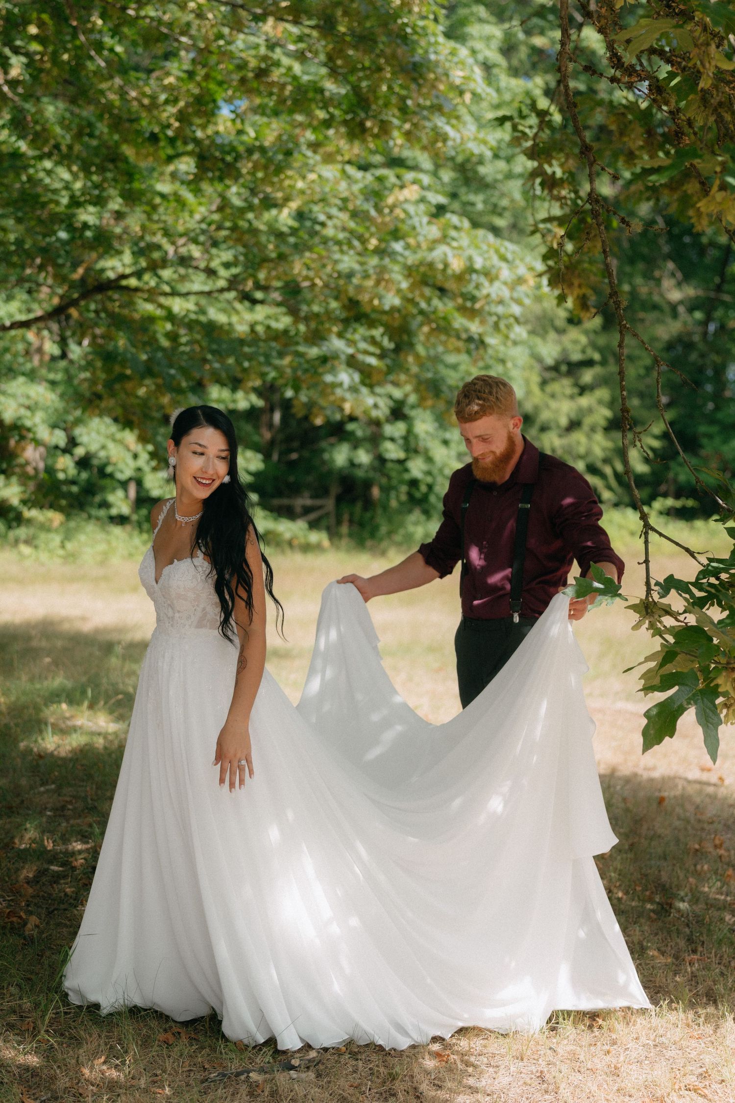 Groom in burgundy shirt arranging bride's white wedding gown train outdoors in a lush green forest setting.