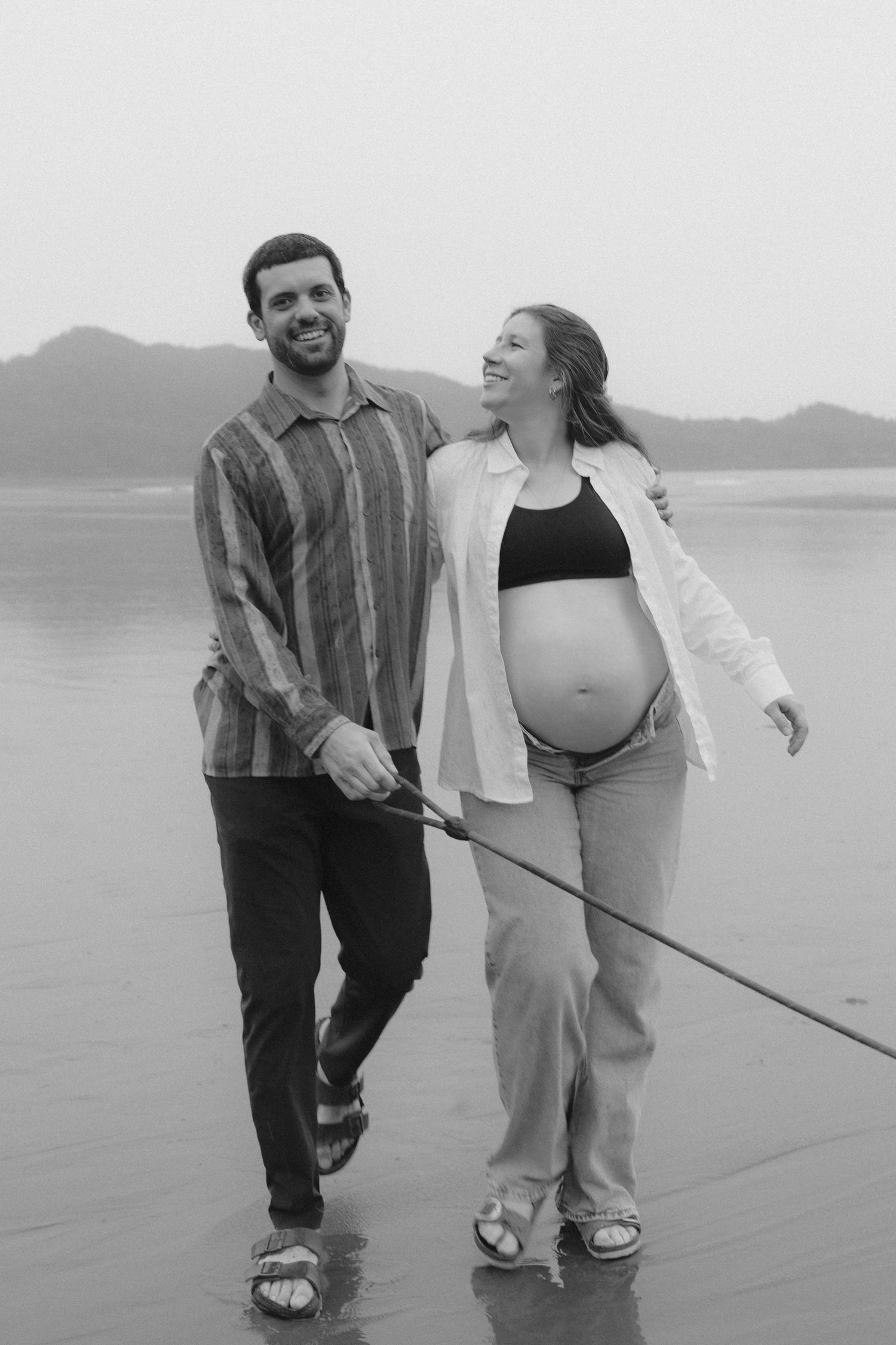 A couple strolls together on a cloudy beach with waves in the background.