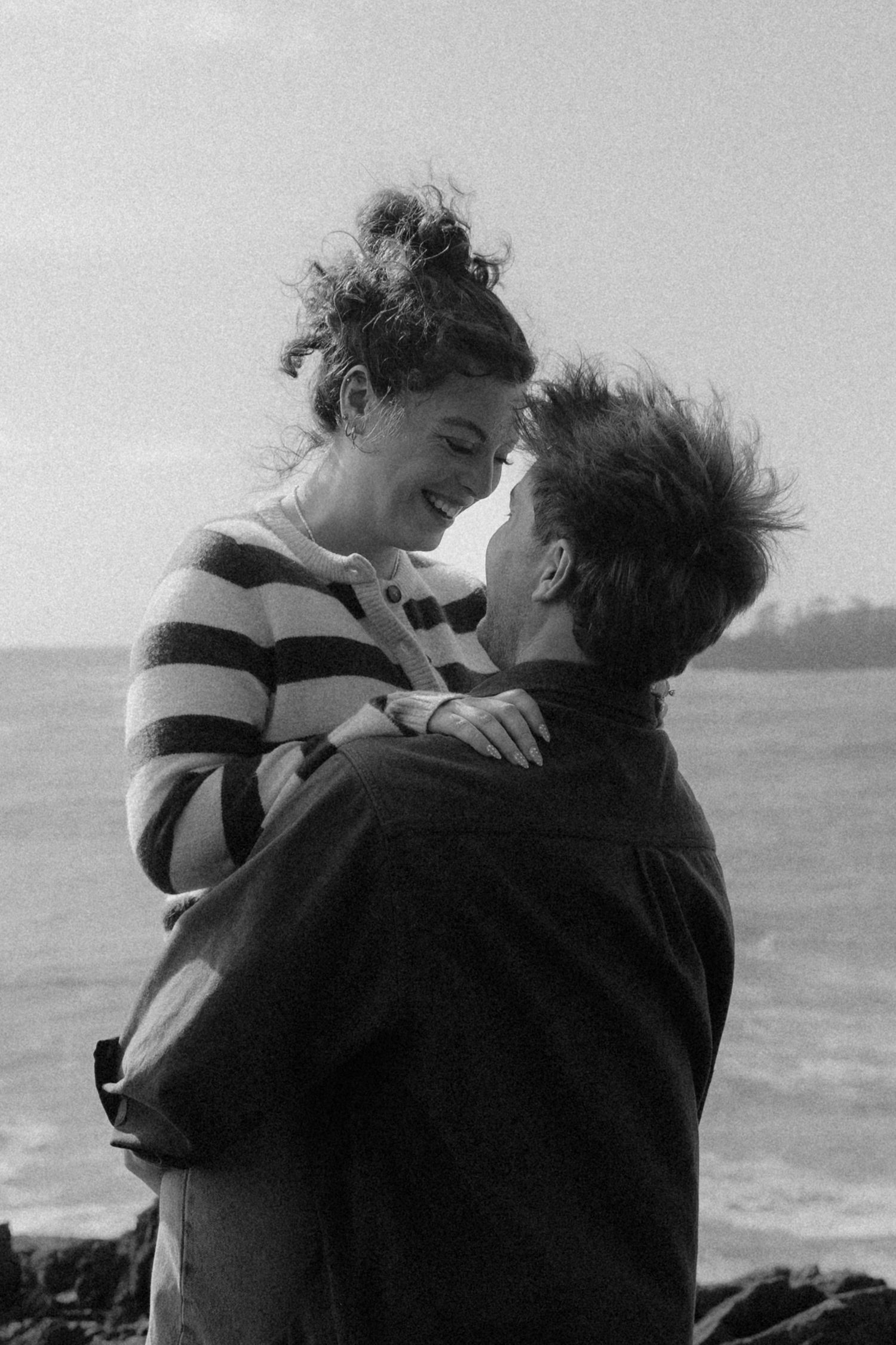 Black and white photo of a joyful couple embracing and smiling at each other by the ocean.