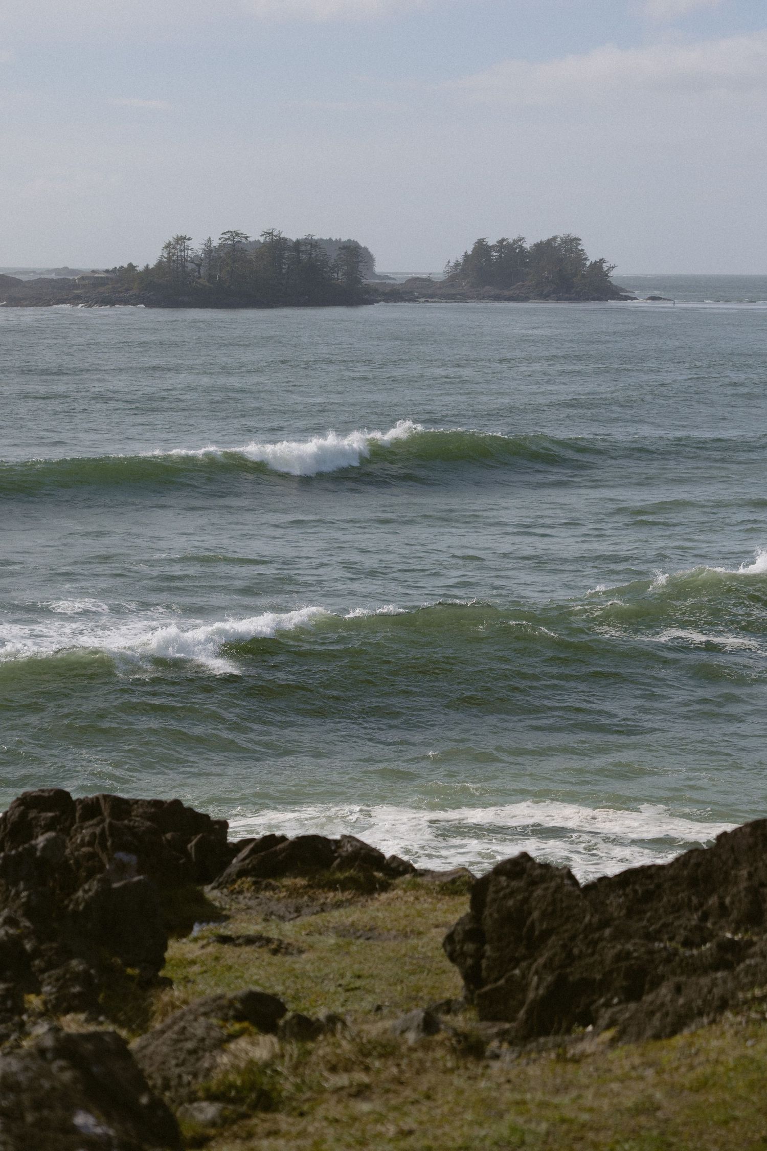 Rocky shoreline with green waves and a tree-covered island in the distance on a cloudy day.