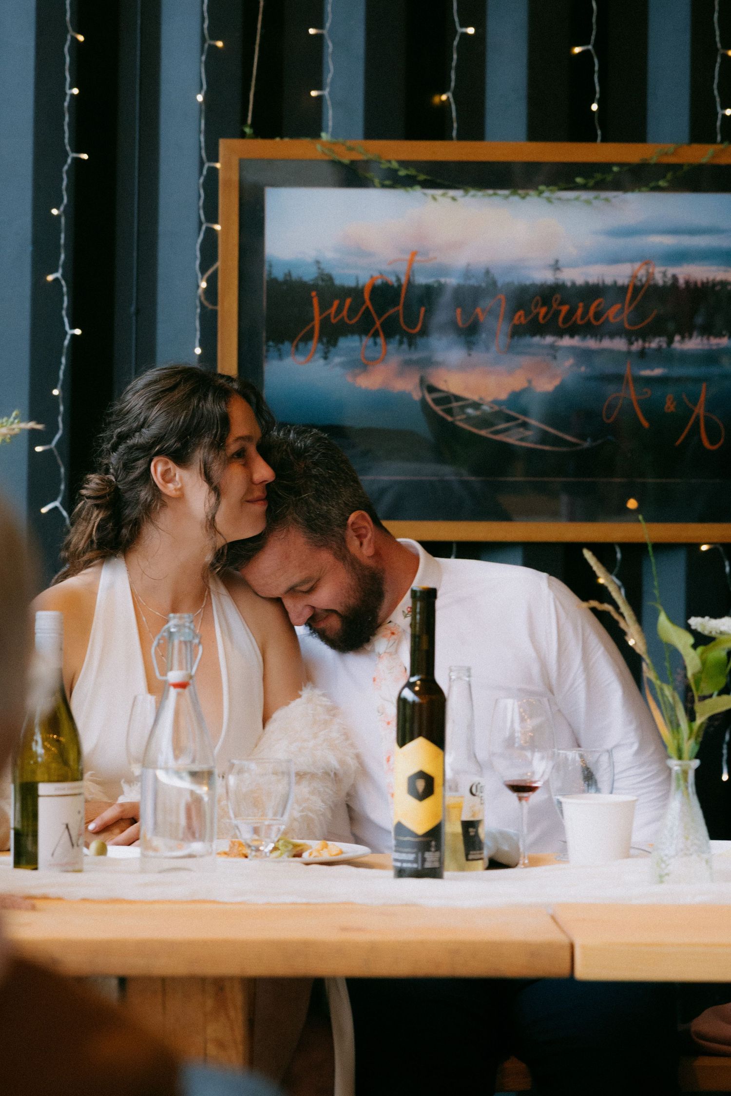 Newlyweds sharing an intimate moment at reception table with just married sign and string lights in background.