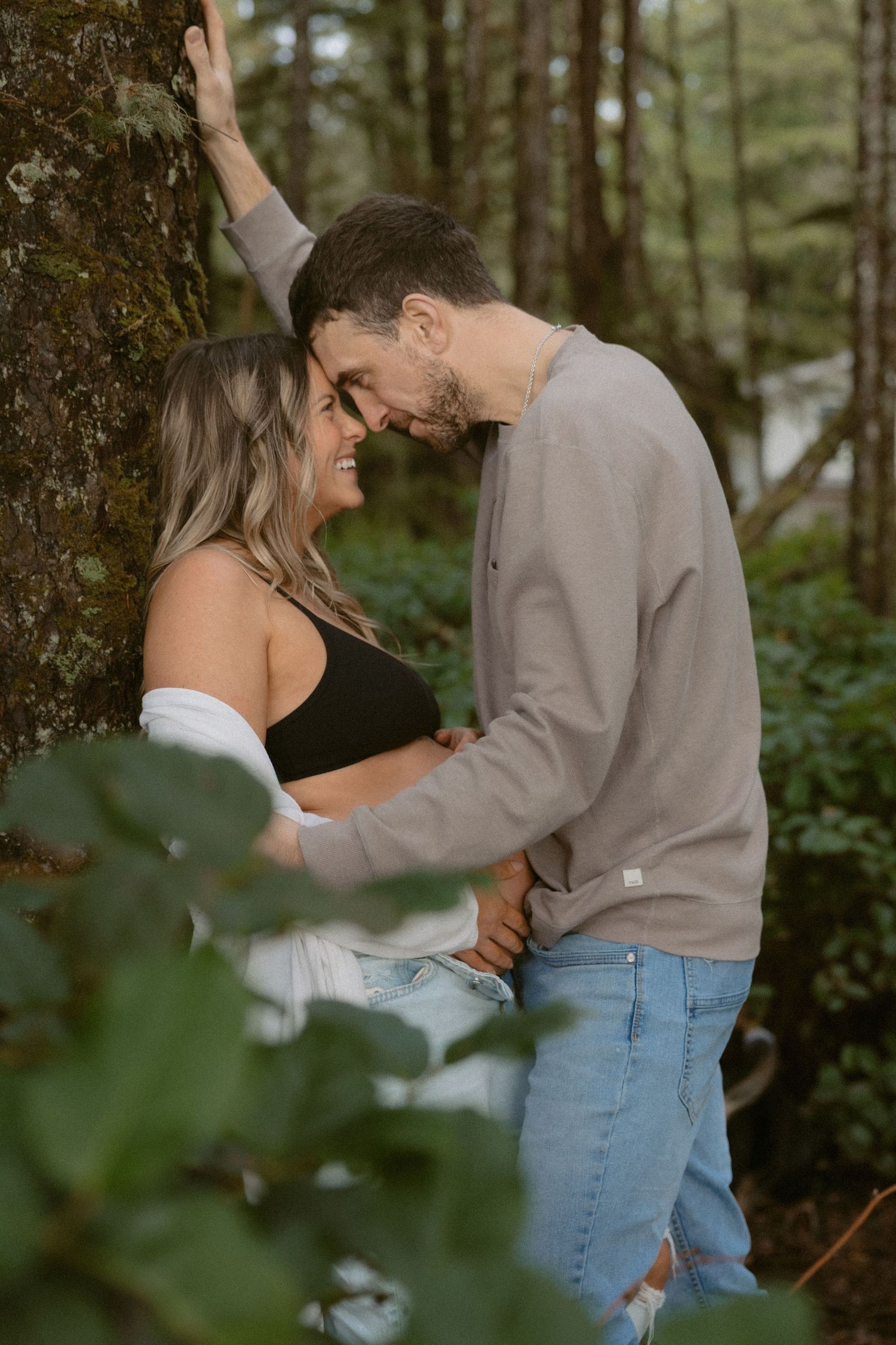 Tofino maternity couple shares an intimate moment against a tree in a lush forest near Chesterman Beach in Tofino