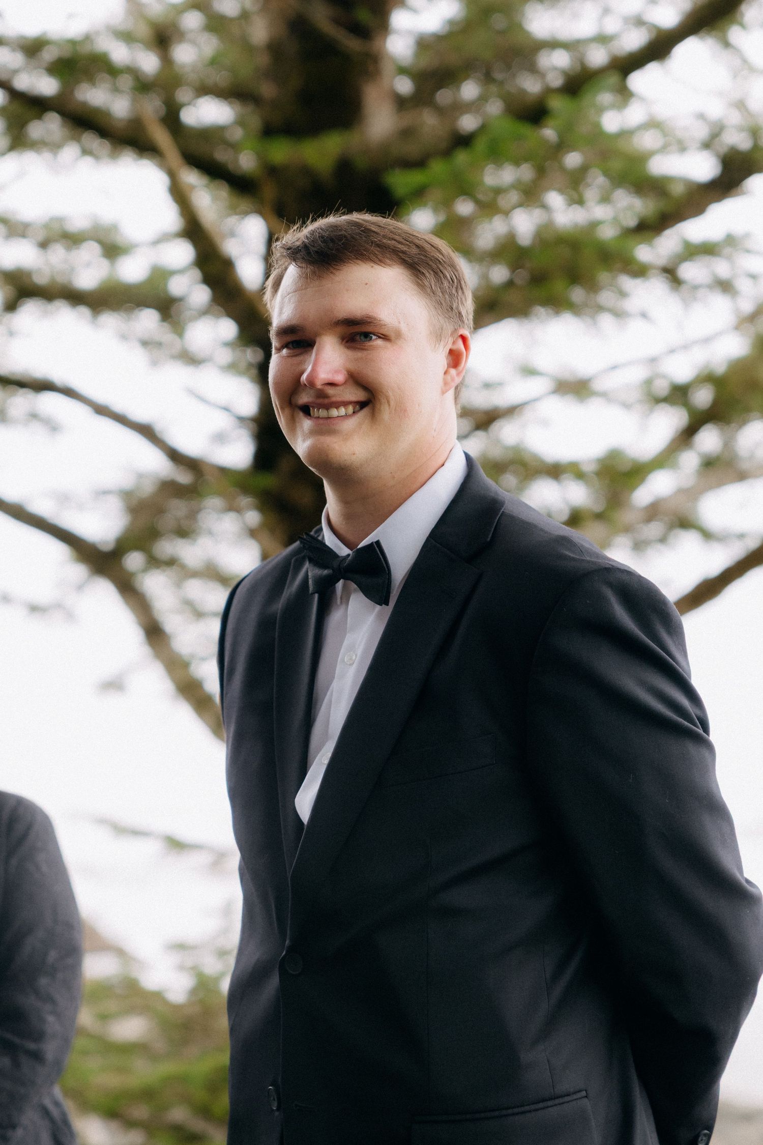 Smiling groom in black tuxedo and bow tie standing outdoors under a tree at a wedding ceremony.