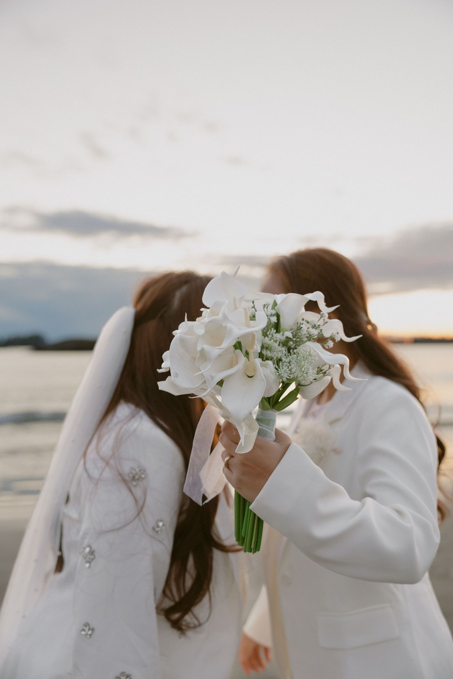 Two people share an intimate moment behind a white bouquet at sunset by the ocean.