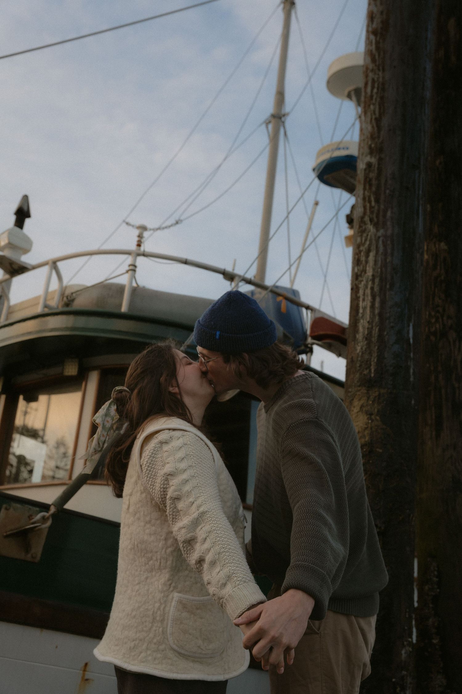 A couple sharing an affectionate moment on a dock with boats and marina buildings behind them.