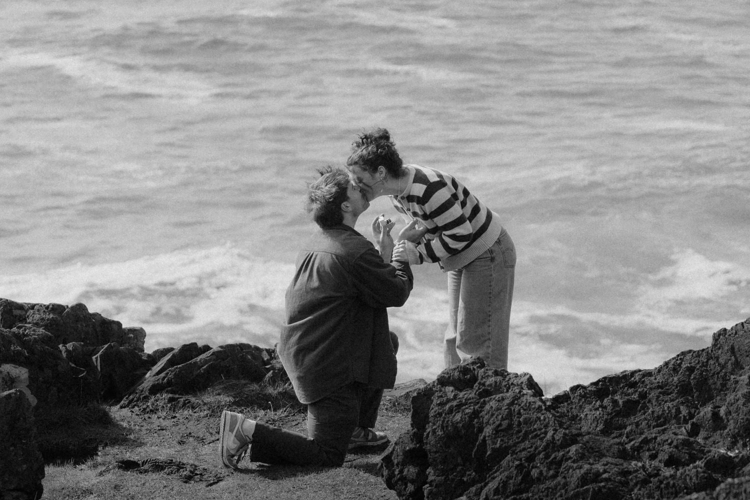 Black and white photo of a person proposing on one knee by rocky ocean shore.