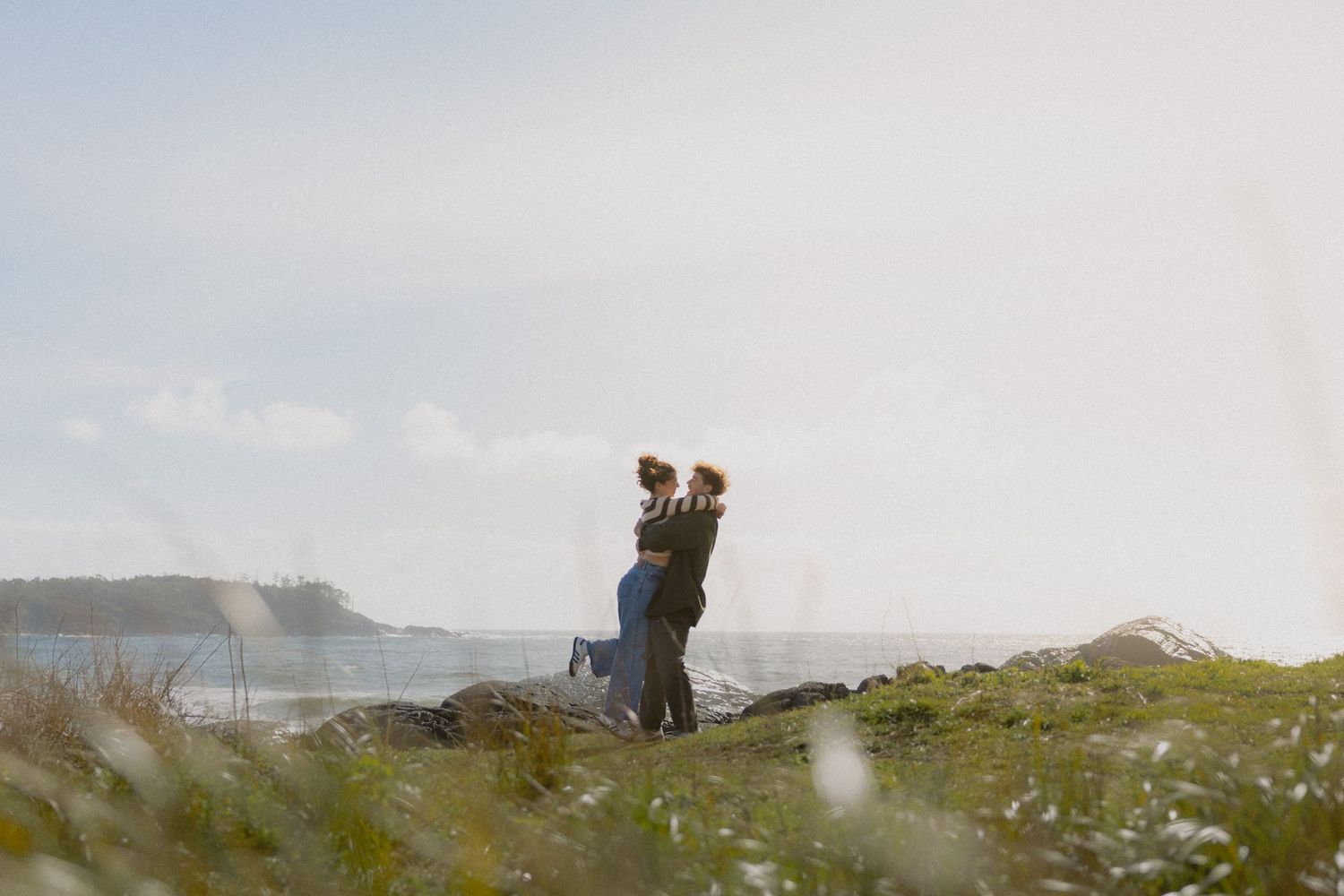 Couple embracing on grassy coastal bluff with dramatic ocean and sky behind them.