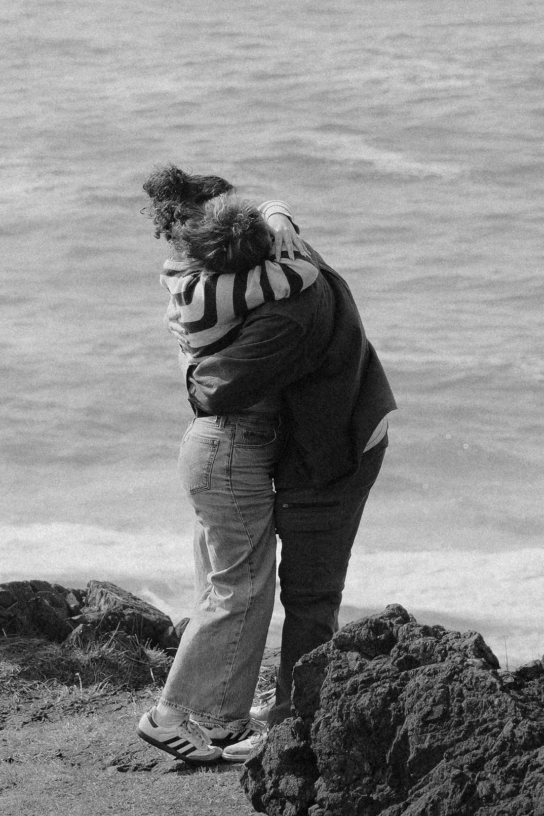 Black and white photo of a couple embracing on rocky coastal cliffs with ocean waves in the background.