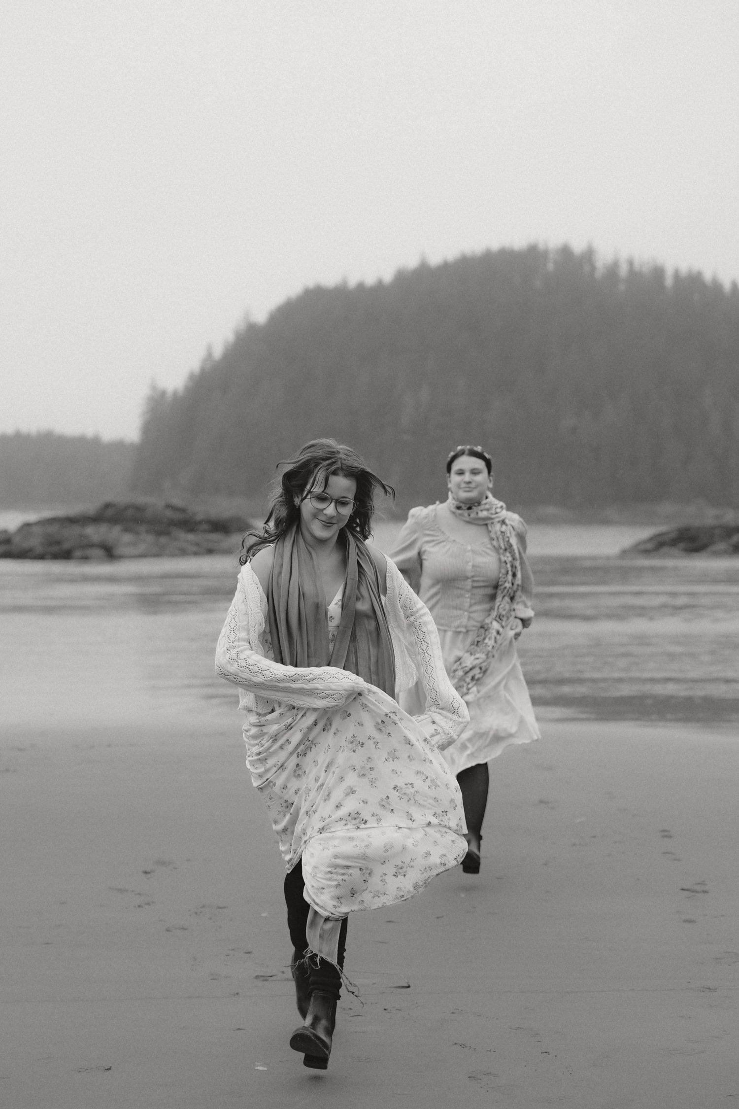 A figure in white lace runs playfully along a misty Pacific Northwest beach with evergreen trees in the background.
