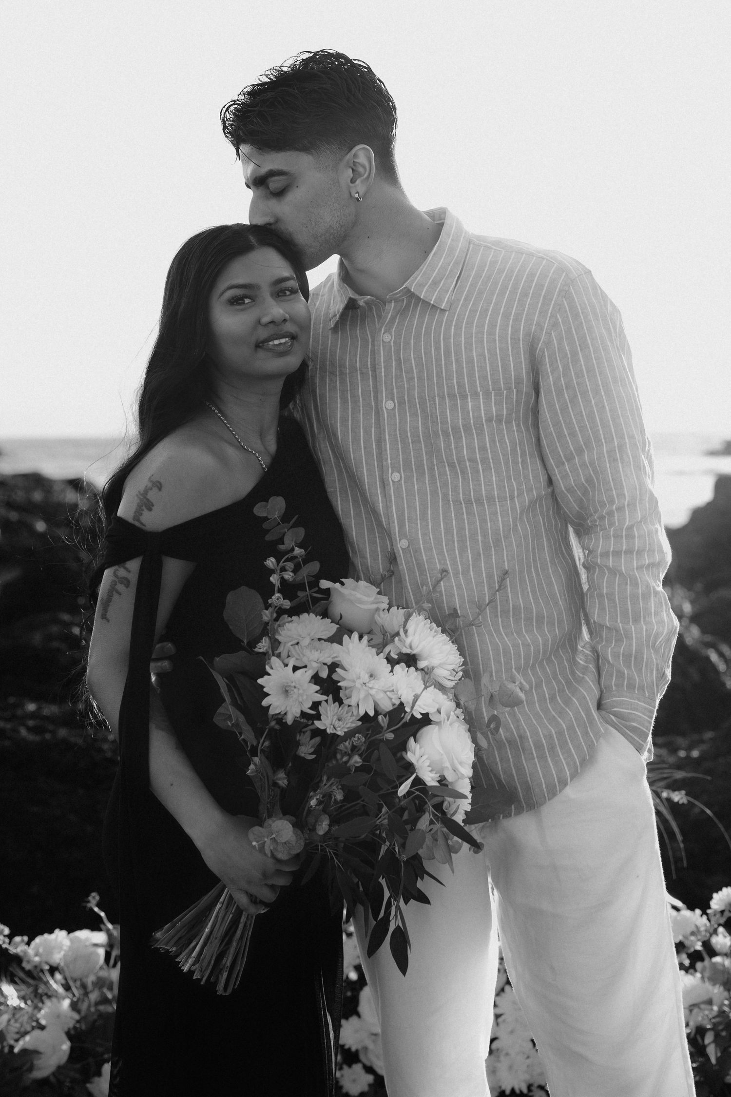 Couple sharing a romantic moment with flowers by the ocean in black and white photography.