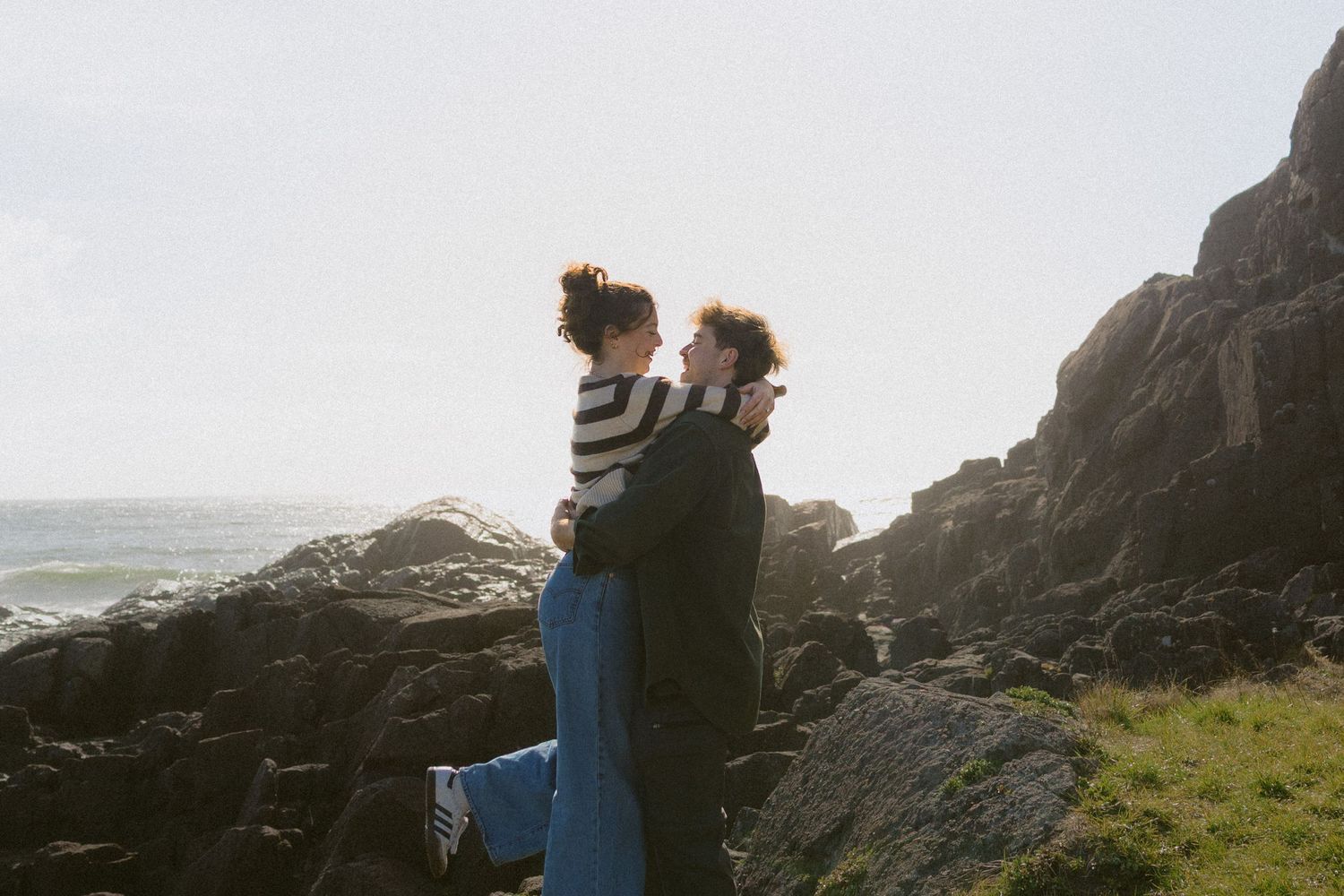 Couple standing on rocky coastal cliffside, man lifting woman in striped top with ocean backdrop.