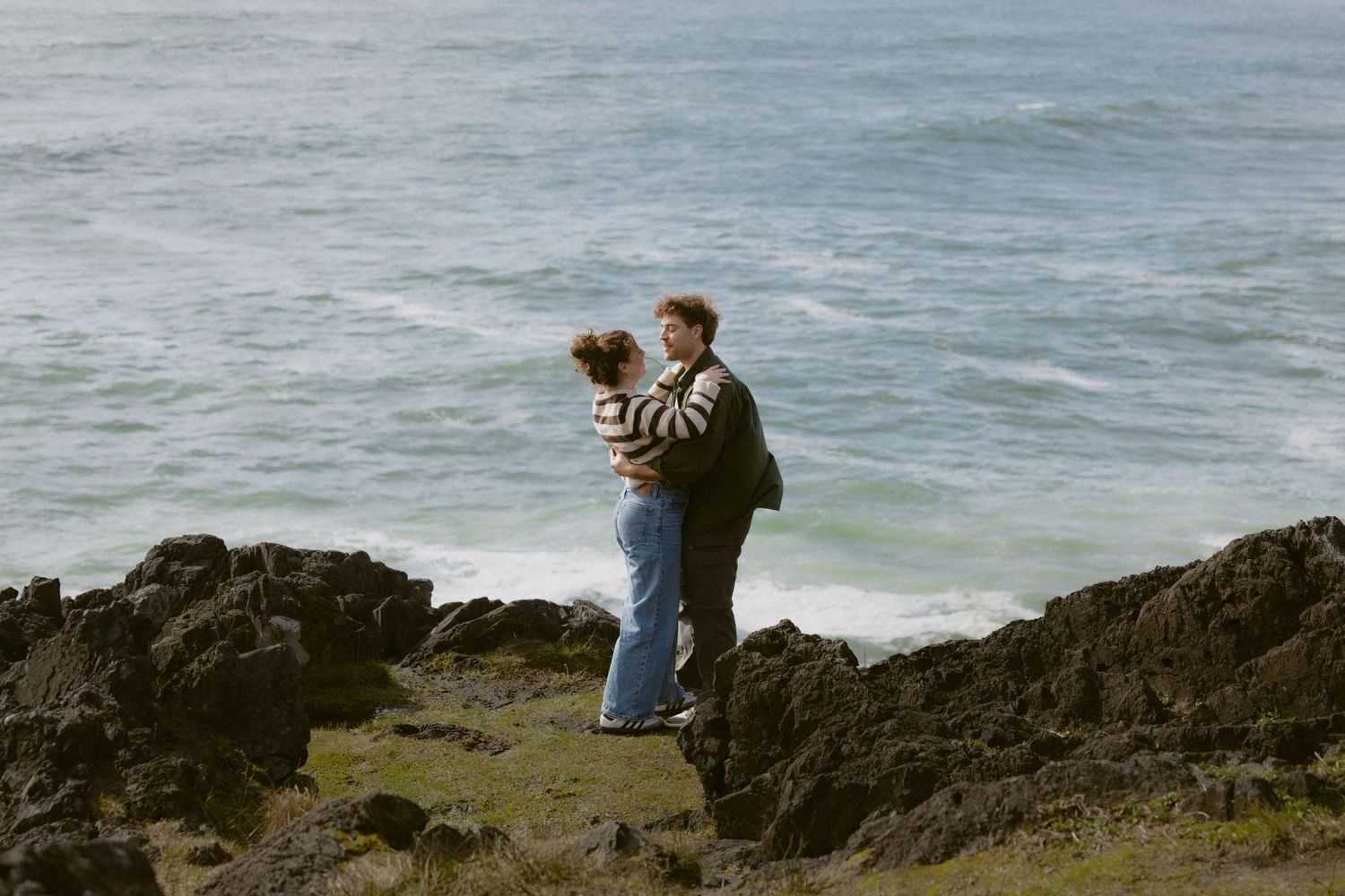 Romantic couple embracing on rugged rocky coast with turquoise ocean waves crashing behind them.