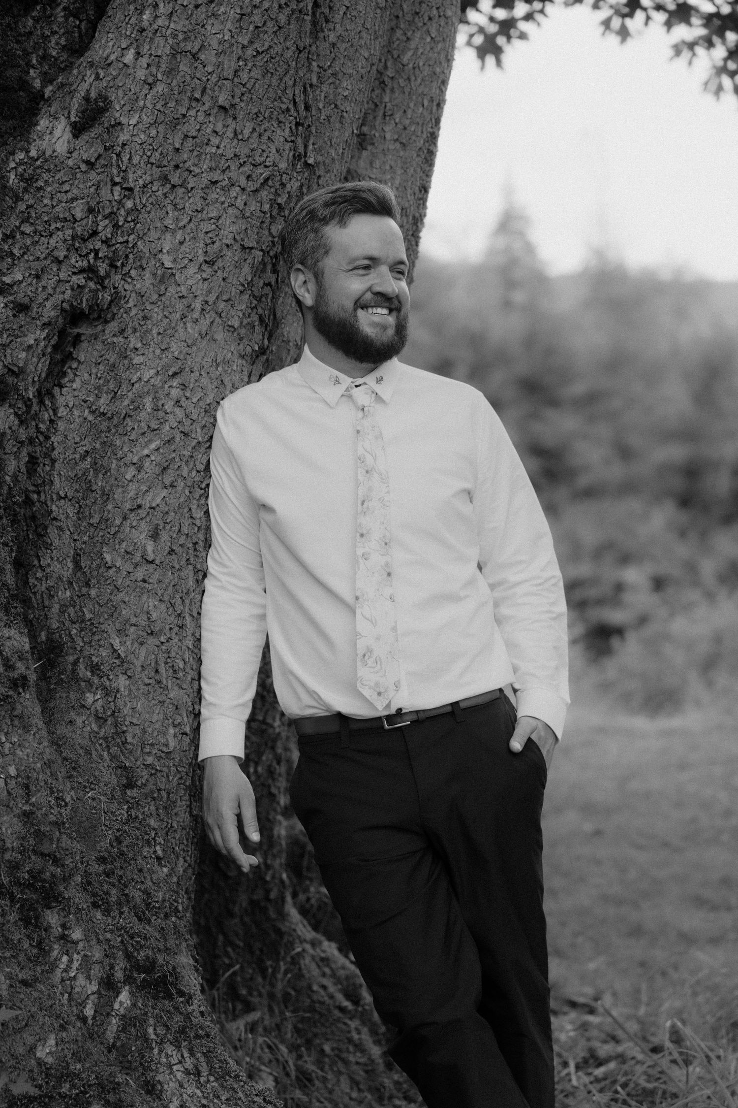Smiling bearded groom in white shirt and floral tie leaning against large tree in black and white outdoor portrait.