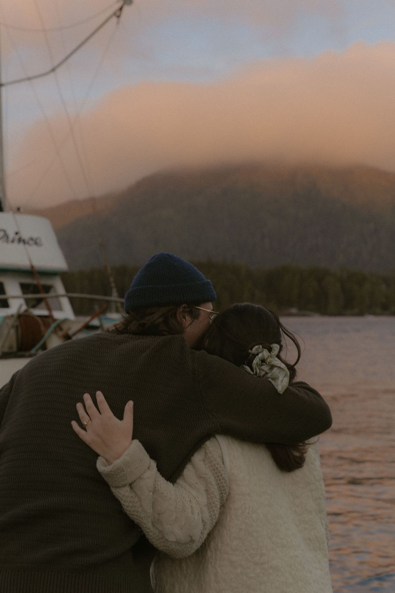 Two people embrace on a dock with mountains and water creating a romantic sunset backdrop.