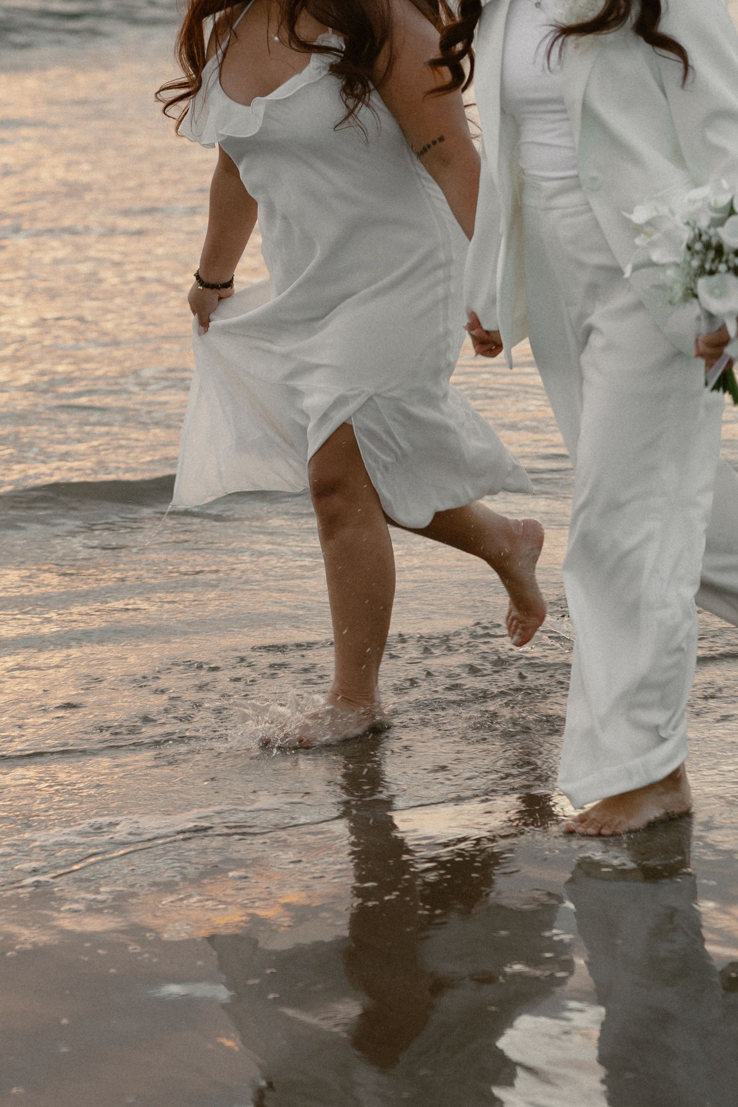 Tofino queer wedding couple walking in shallow water at the beach wearing wedding dresses