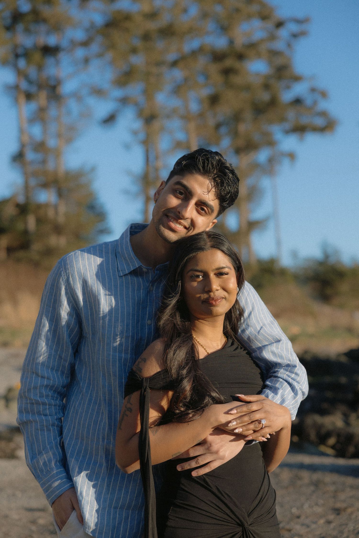 Couple embracing outdoors at golden hour, man in blue shirt hugging woman in black dress with trees behind.