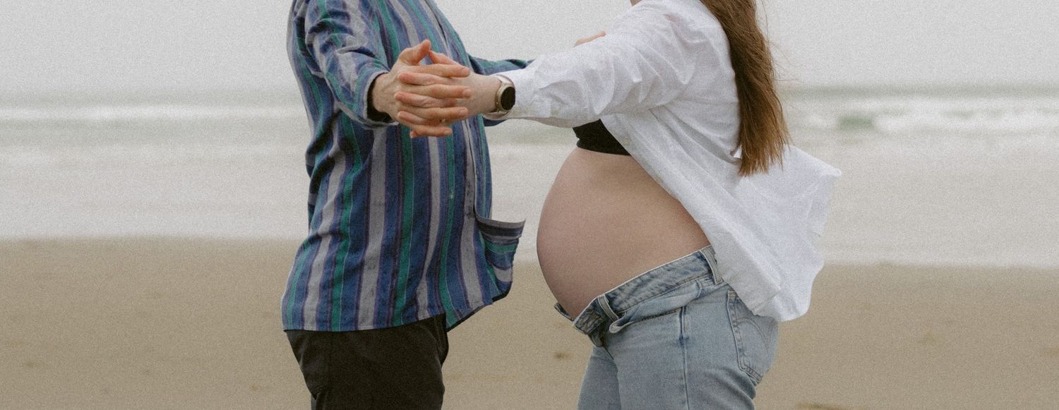 Couple on beach, pregnant woman in white shirt and jeans with partner in striped shirt touching her baby bump.