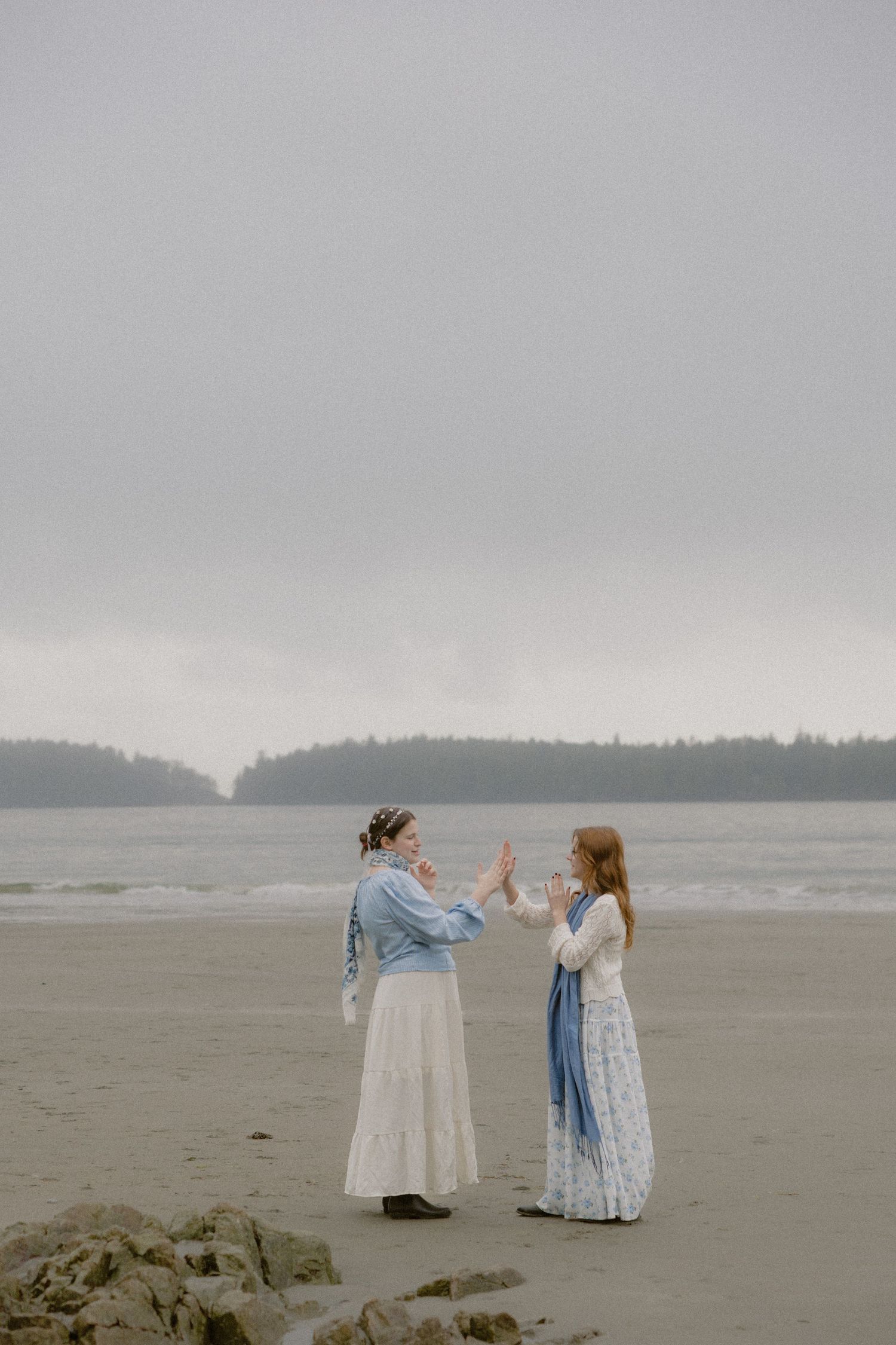 People in vintage-inspired outfits dance together on a wide sandy beach under overcast skies.