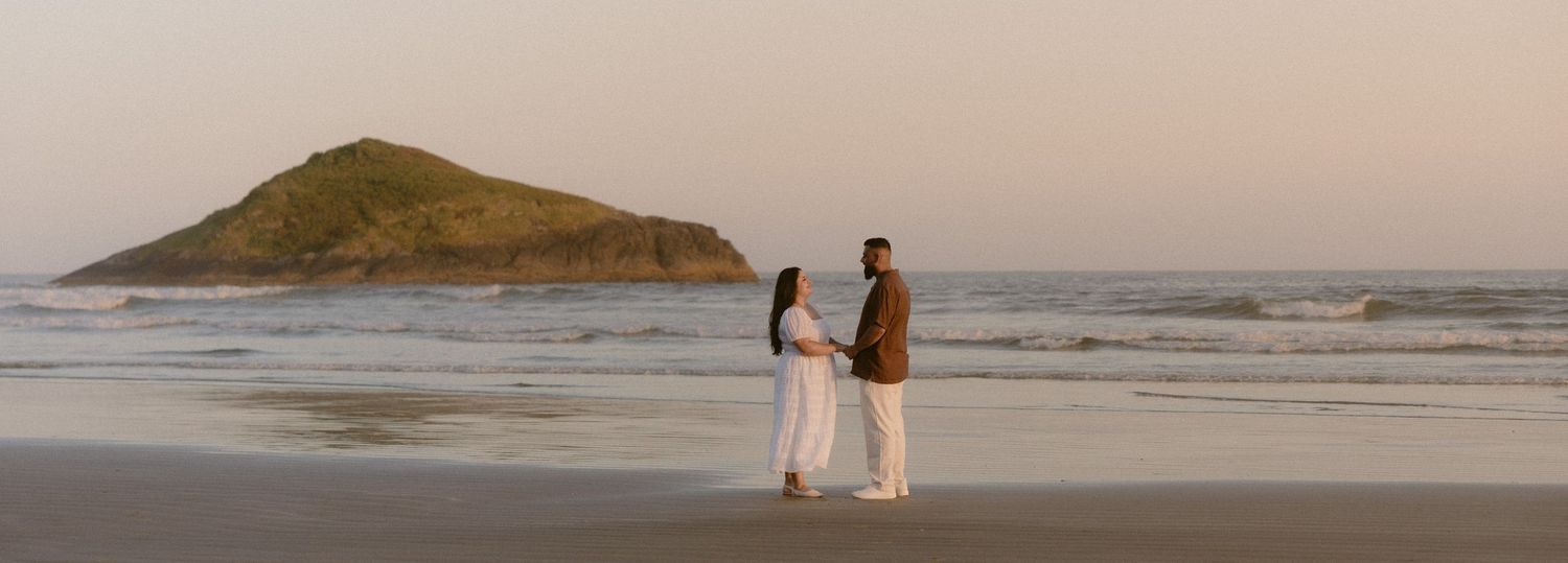 Couple holding hands on a sandy beach at sunset with a scenic green hill and ocean waves in the background.