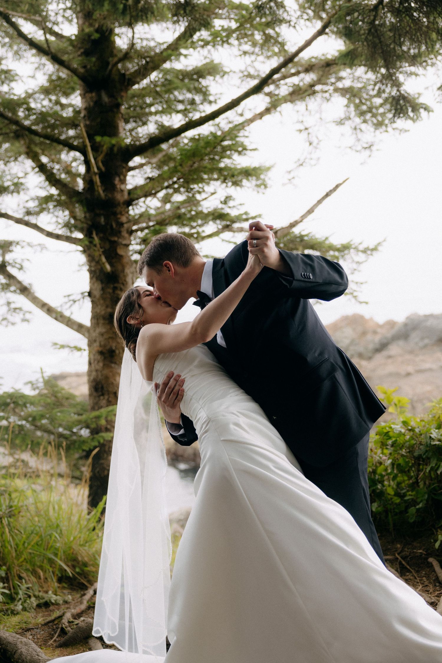 Bride and groom sharing a romantic dip kiss outdoors near trees during their wedding celebration.