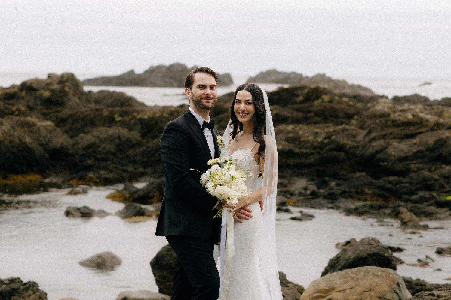 Bride and groom posing on rocky beach, bride holding white bouquet in elegant gown, groom in black tuxedo.